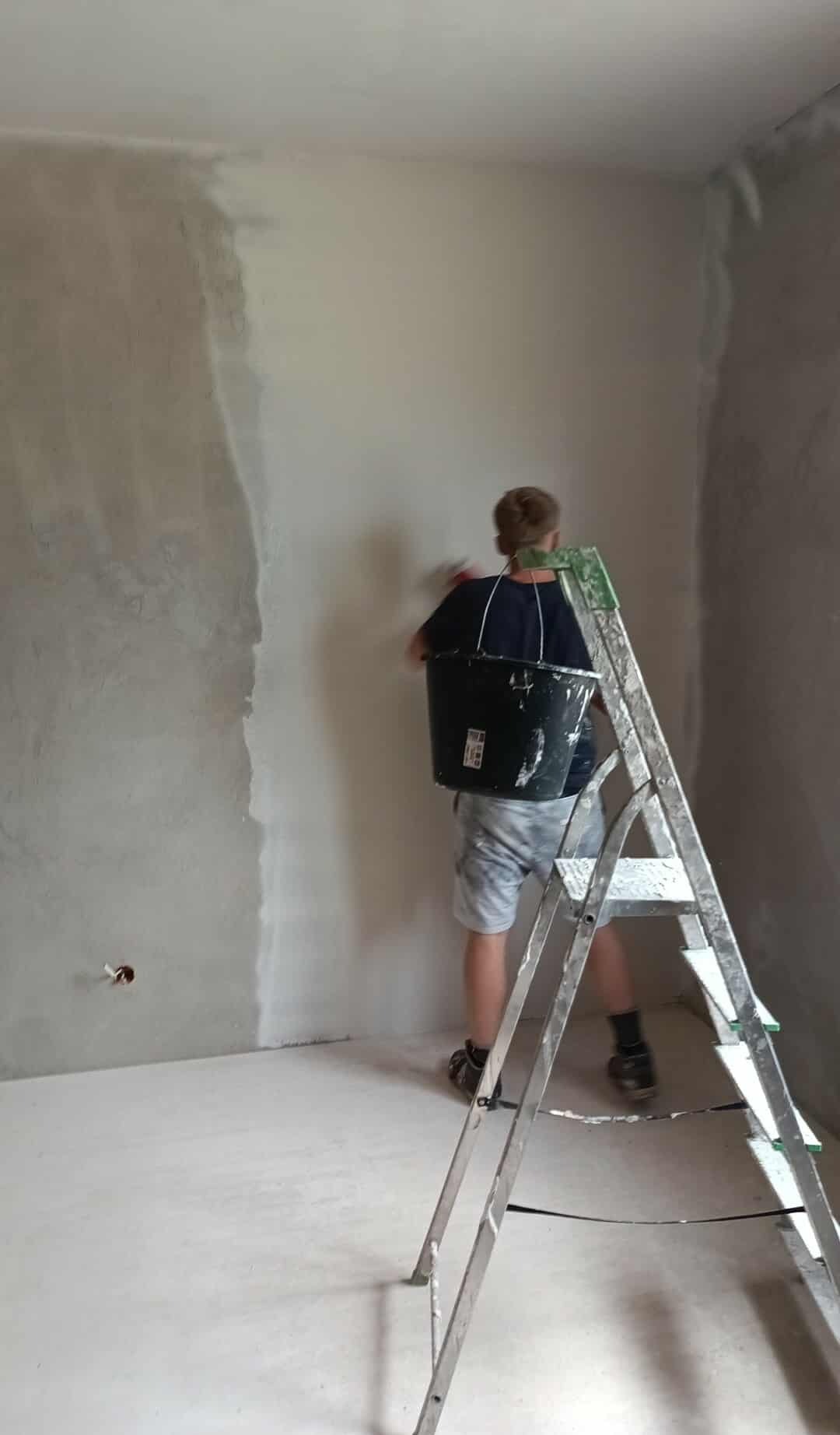 Mężczyzna w trakcie szpachlowania ściany w rogu pokoju, z wiadrem zawieszonym na plecach i drabiną obok. Widoczny fragment ściany przed i po szpachlowaniu.