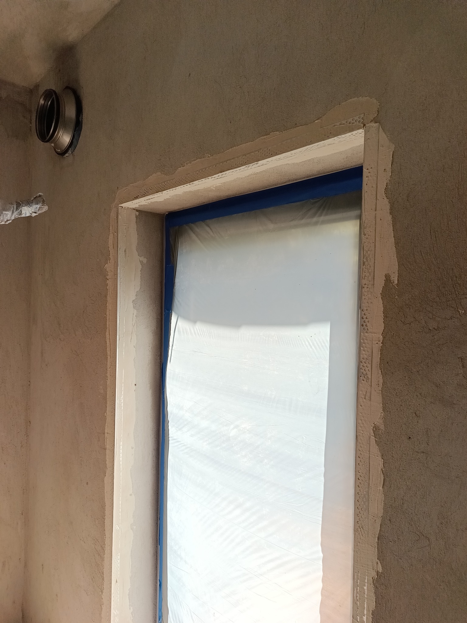 Ściana w trakcie szpachlowania przy oknie, zabezpieczonym folią i taśmą. Widoczny fragment instalacji wentylacyjnej. Nierówna powierzchnia ściany przygotowana do dalszych prac.