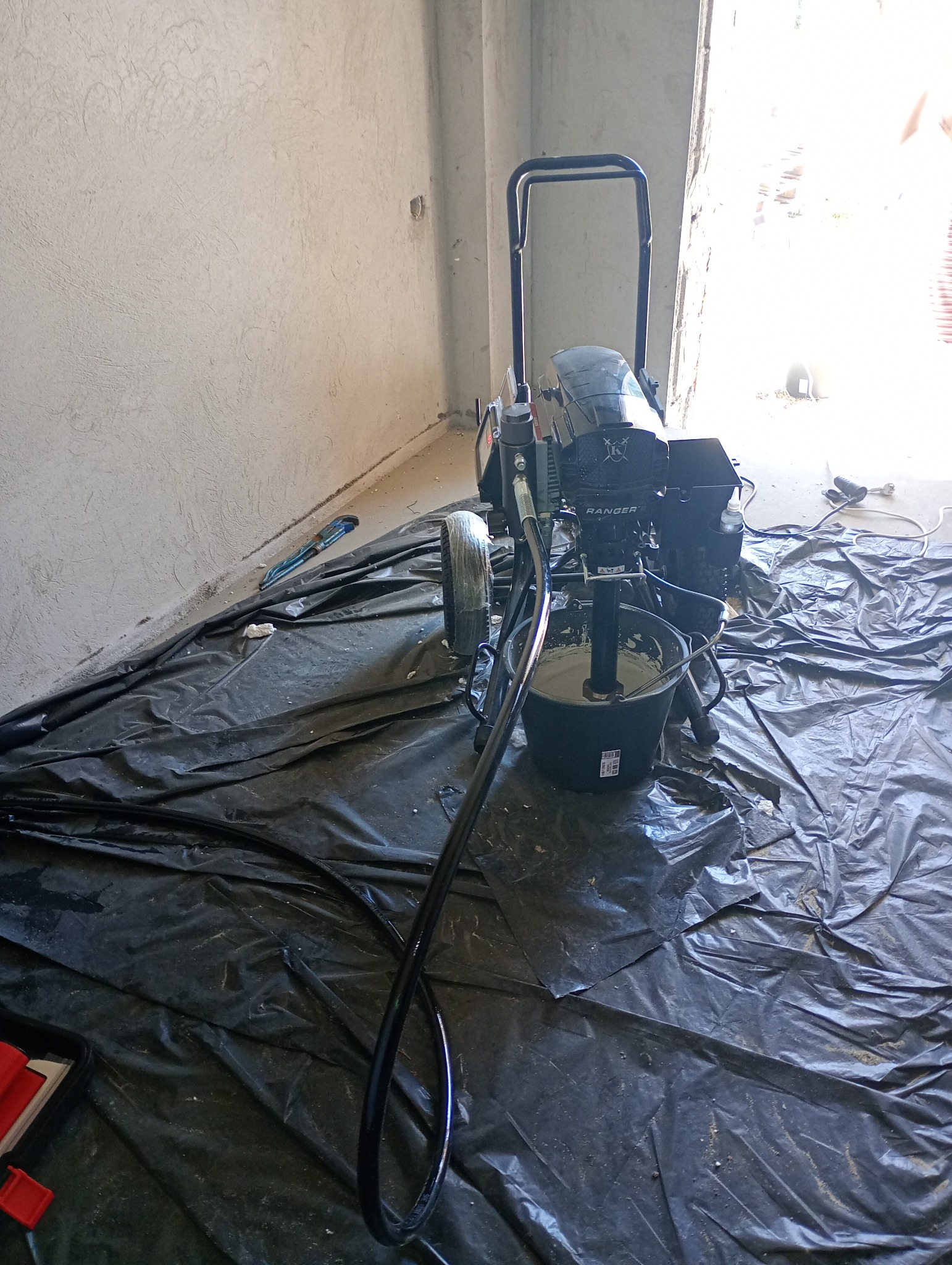 Agregat malarski Ranger z czarnym wężem i wiadrem z farbą stoi na folii ochronnej w rogu pokoju ze ścianami po szpachlowaniu. W tle widoczne okno.