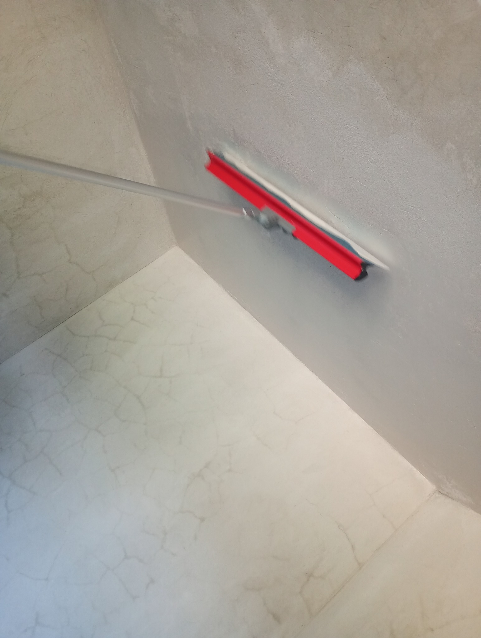 Nakładanie gładzi na ścianę w rogu pomieszczenia za pomocą szpachli z długim uchwytem. Widoczne delikatne wzory na ścianie i podłodze.