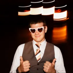 Uśmiechnięty mężczyzna w okularach w kształcie serc, kamizelce i krawacie, pokazuje kciuki w górę na ciemnym tle z rozmytymi światłami.