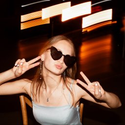 KK Pracownia Fotograficzna Karina Kolenda - Młoda kobieta w okularach w kształcie serc pozuje z gestem zwycięstwa na tle rozmytych świateł, siedząc na krześle. Ubrana w sukienkę na ramiączkach i biżuterię.