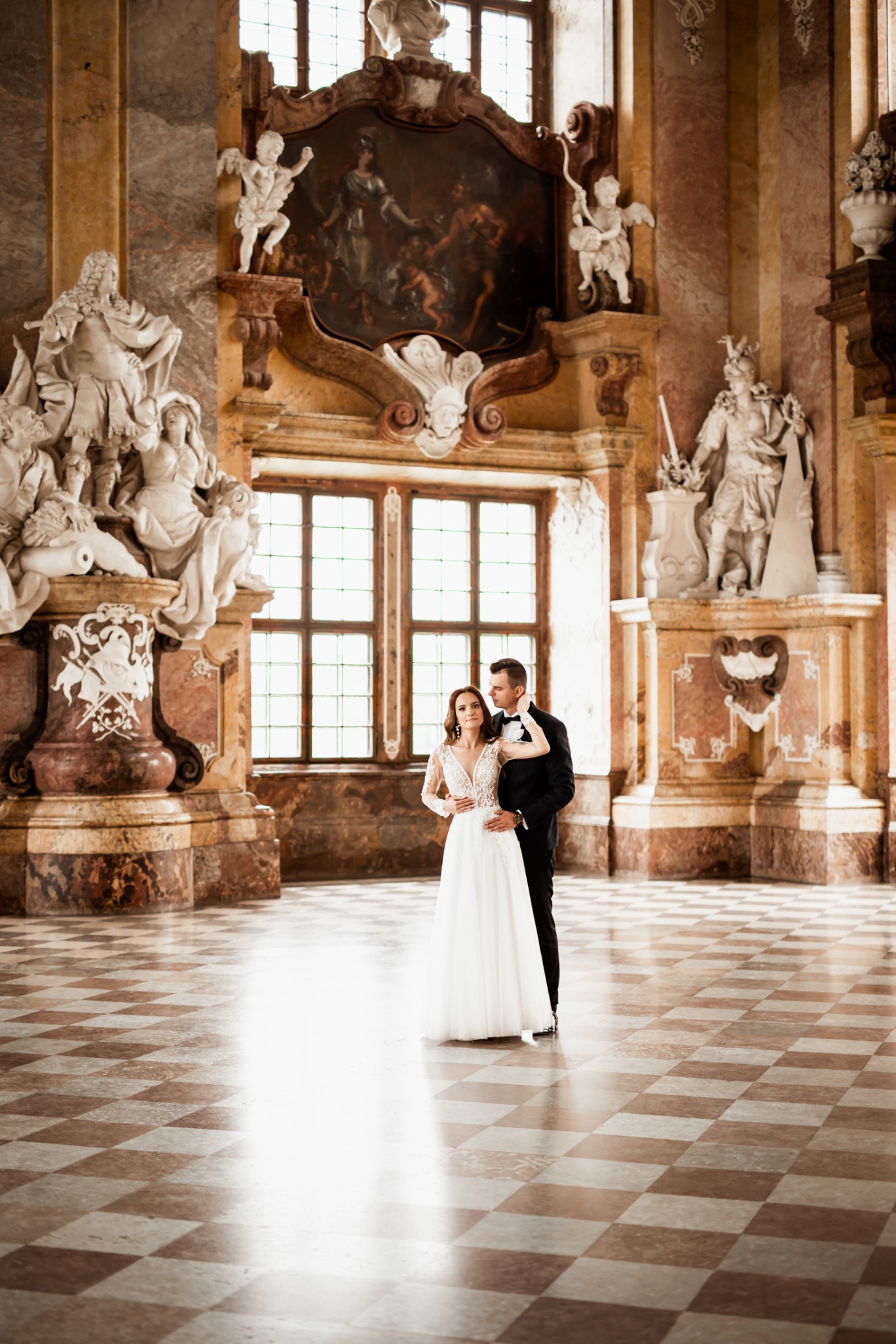 Elegancka para młoda w barokowym wnętrzu: on w smokingu, ona w koronkowej sukni. Marmurowe posadzki i bogate zdobienia podkreślają uroczysty charakter sesji.