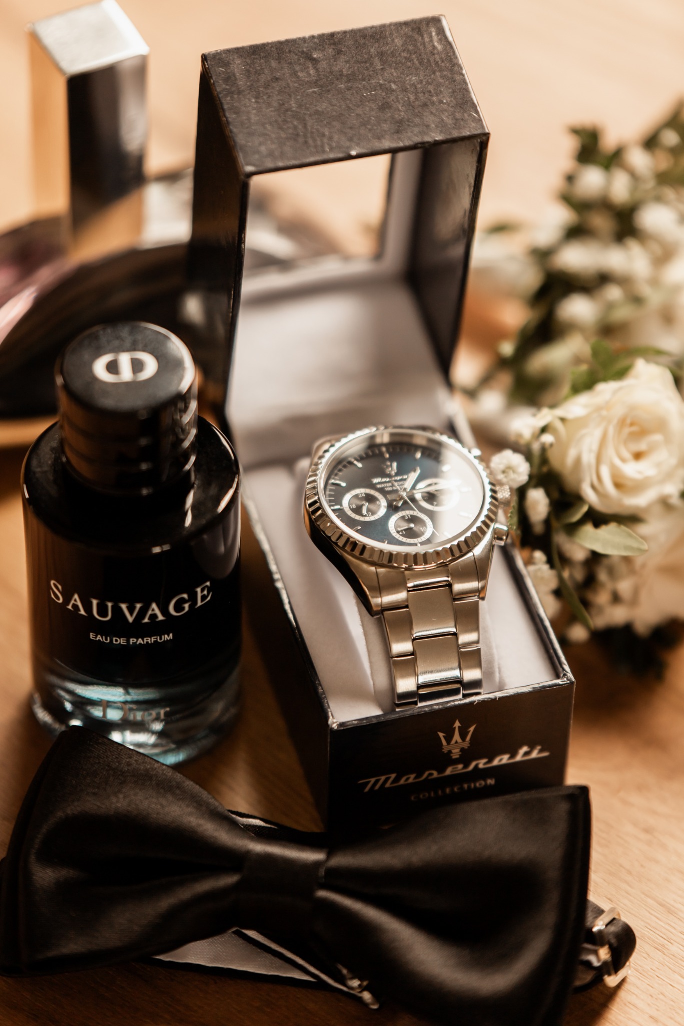 Elegancki zestaw ślubny: zegarek Maserati w pudełku, perfumy Sauvage, czarna mucha i bukiet białych róż na drewnianym stole. Detale przygotowań do ślubu.