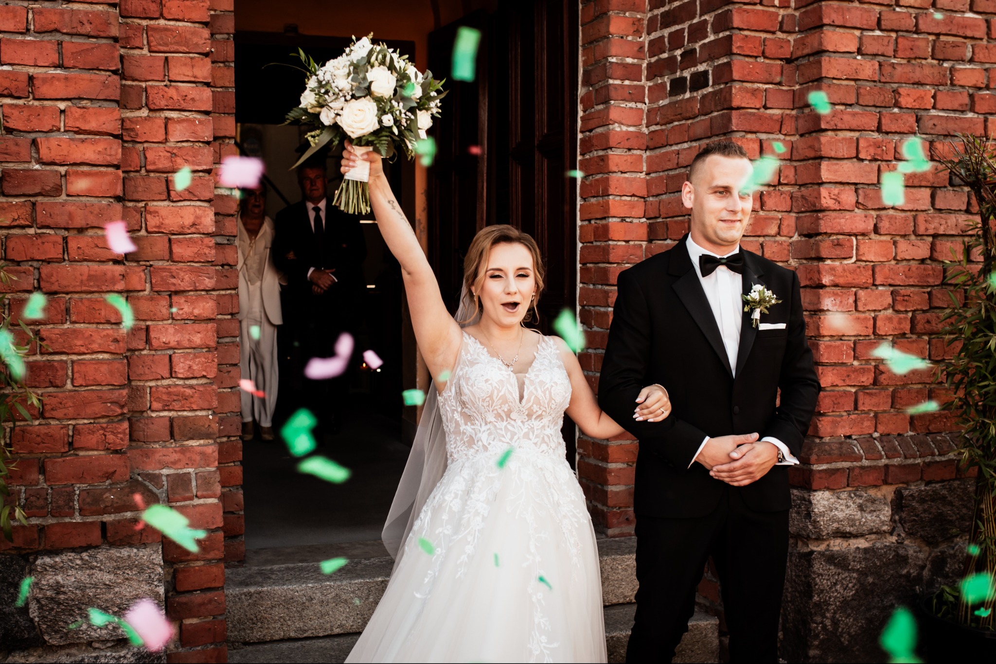 Uśmiechnięta panna młoda z bukietem i pan młody w smokingu wychodzą z kościoła, obsypywani konfetti na tle ceglanej ściany.