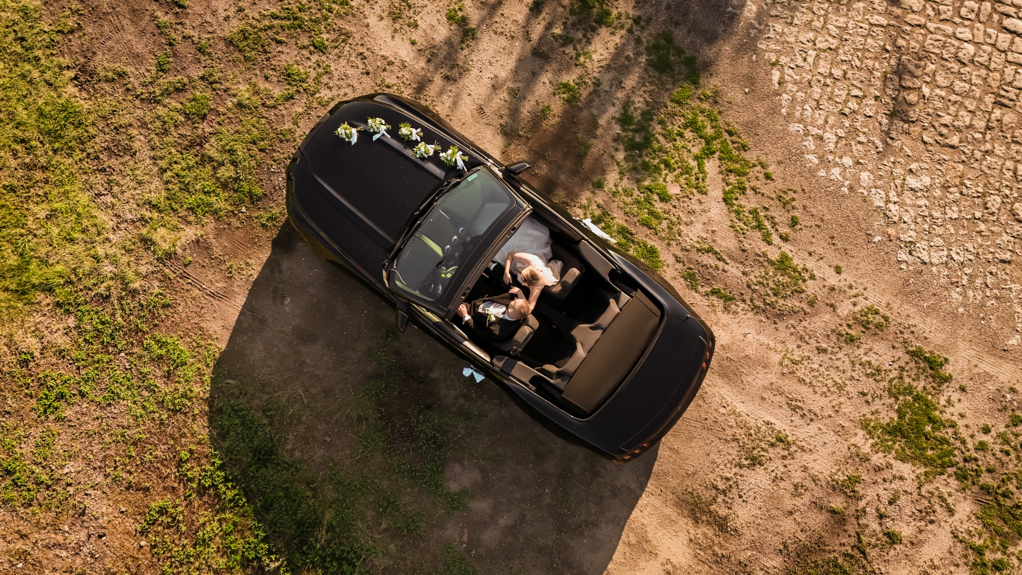 Czarny kabriolet z kwiatową dekoracją na masce, widziany z góry. W środku para młoda w strojach ślubnych, na tle ziemistej drogi i zieleni.