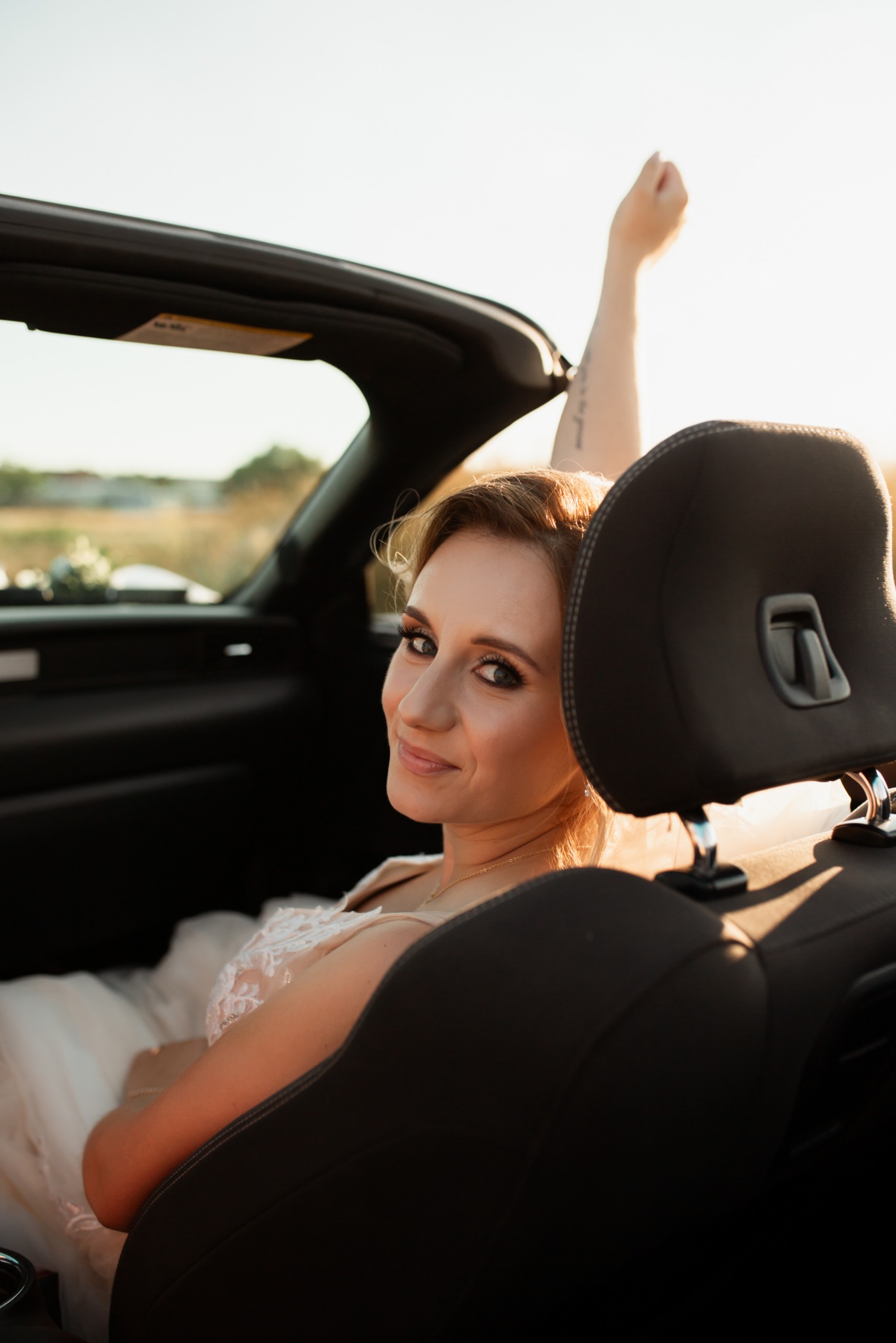 Uśmiechnięta panna młoda w sukni z koronką, siedząca w kabriolecie z uniesioną ręką na tle złotego pola, sfotografowana zza ramienia fotela.