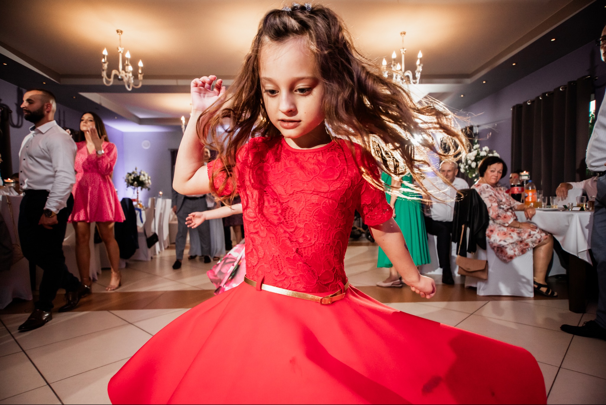 Dziewczynka w czerwonej sukience tańczy na parkiecie podczas imprezy. W tle widoczni goście i dekoracje sali balowej z żyrandolami.