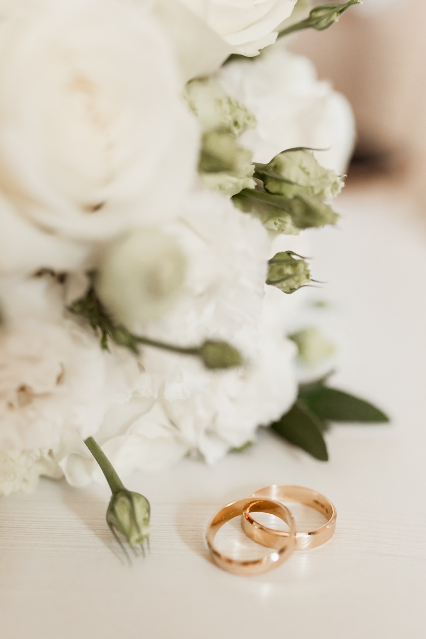 Dwa złote obrączki ślubne na białym tle, obok bukiet białych kwiatów. Delikatne światło podkreśla detale. Ujęcie z bliska, skupione na symbolice związku.