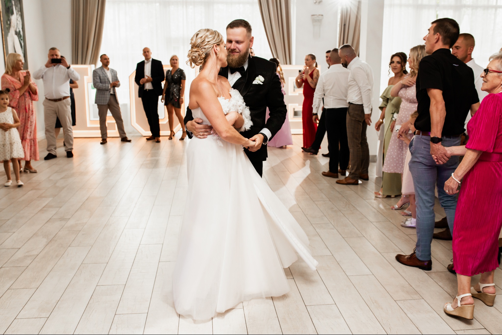 Para młoda w tańcu podczas wesela w Kąkolewie. Panna młoda w białej sukni, pan młody w czarnym smokingu. Goście weselni obserwują i robią zdjęcia.