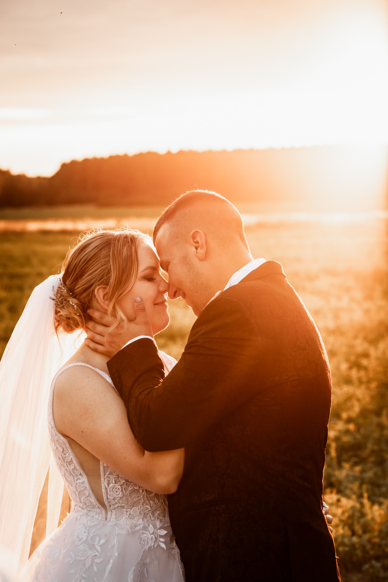 Złocista sesja ślubna w plenerze: para młoda w objęciach, twarze blisko, zachód słońca w tle, delikatny welon panny młodej i ciemny garnitur pana młodego.