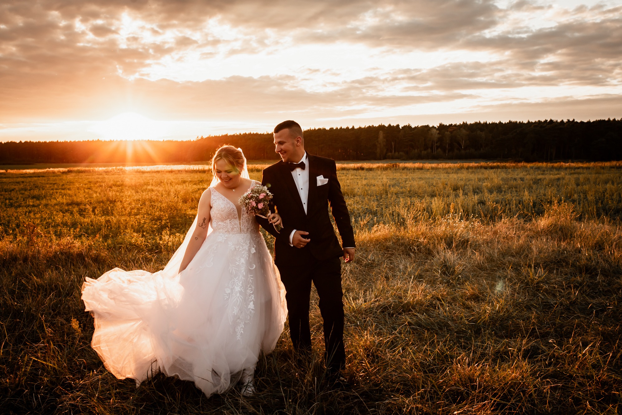 Para młoda spaceruje po łące podczas zachodu słońca. Panna młoda w sukni z trenem, pan młody w smokingu. Romantyczna sceneria z ciepłym światłem.