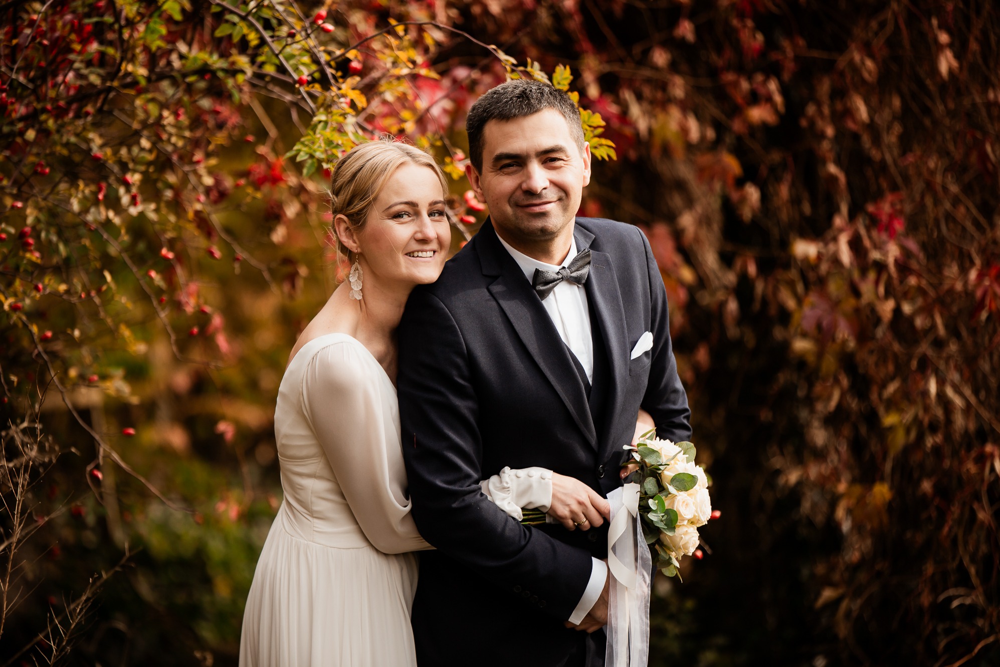 Para młoda w jesiennej scenerii: panna młoda w sukni ivory opiera się o pana młodego w czarnym smokingu z bukietem białych róż. Tło stanowią krzewy z czerwonymi owocami.