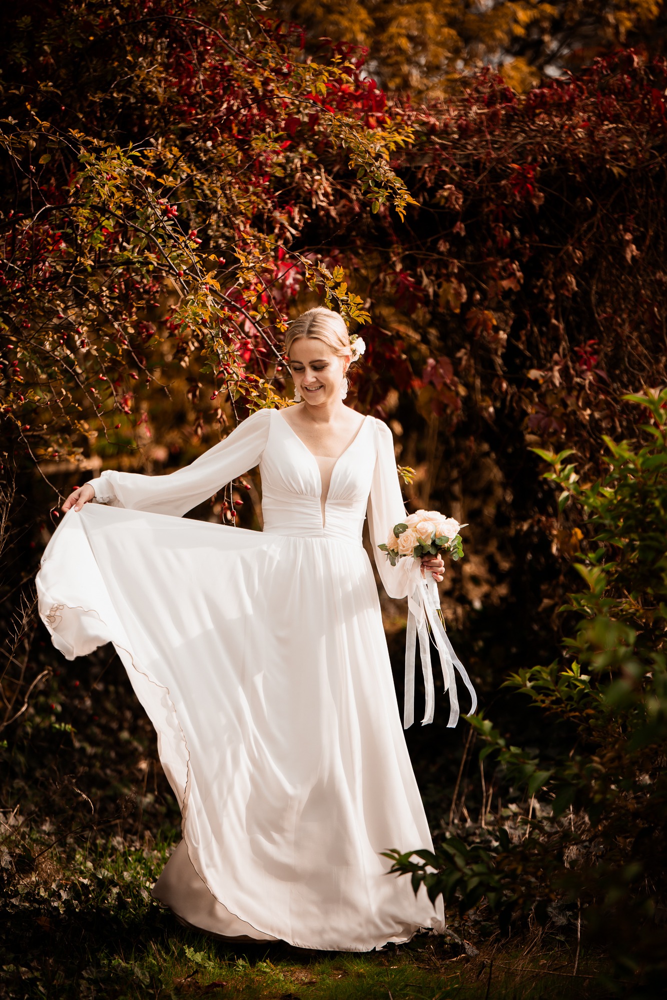 Uśmiechnięta panna młoda w zwiewnej sukni, trzyma bukiet w jesiennej scenerii. Romantyczna sesja ślubna w plenerze.