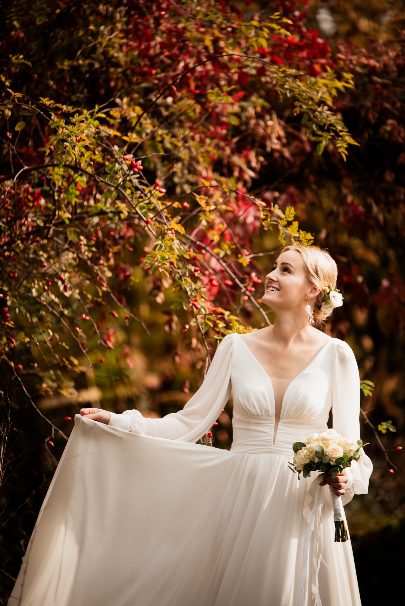 Panna młoda w białej sukni z bukietem, uśmiechnięta, patrzy w górę na tle drzewa z czerwonymi liśćmi. Subtelne oświetlenie podkreśla elegancję i naturalność.