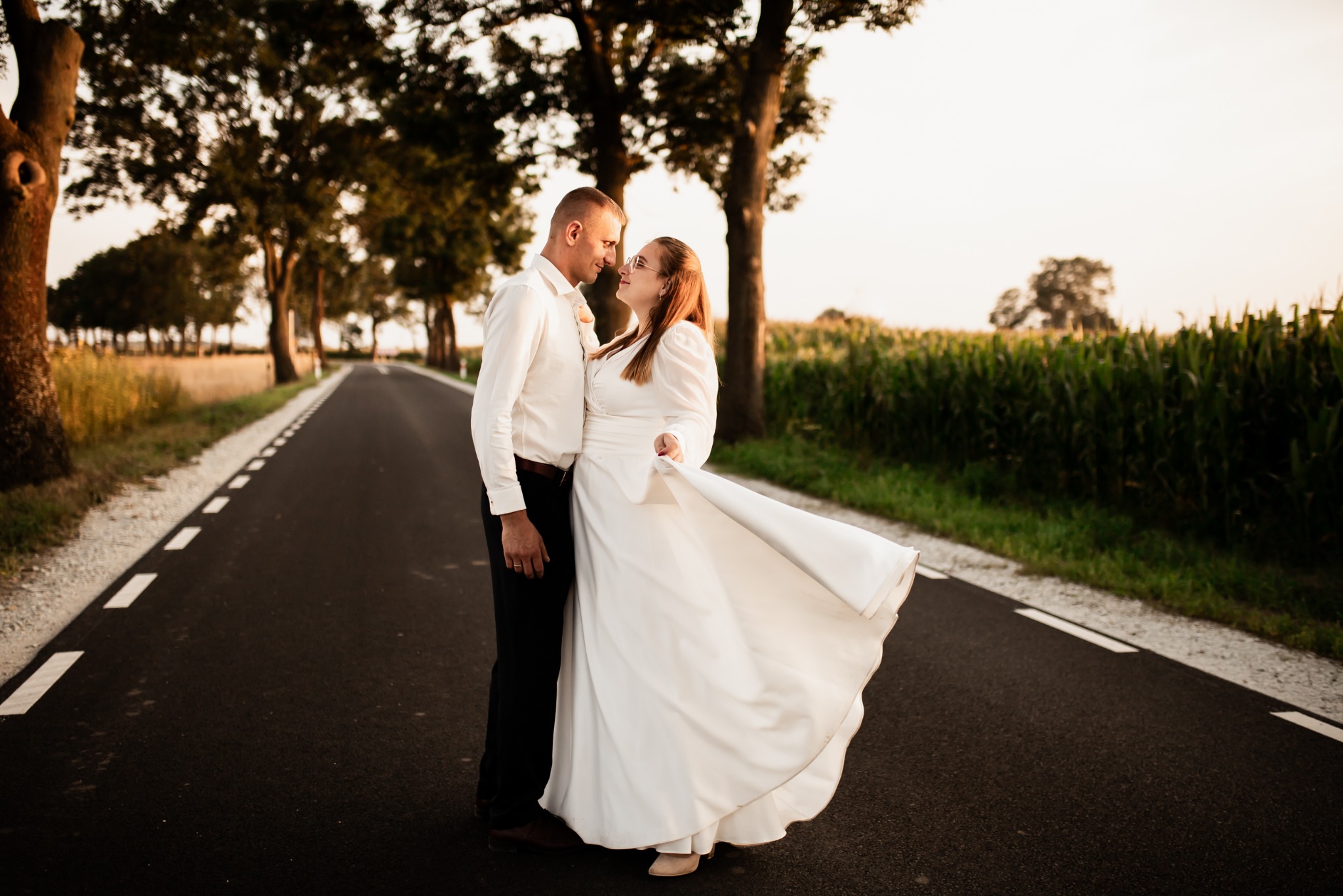 Para młoda na drodze otoczonej drzewami i polem kukurydzy. Panna młoda w białej sukni z podmuchem wiatru. Delikatne, ciepłe światło zachodzącego słońca.