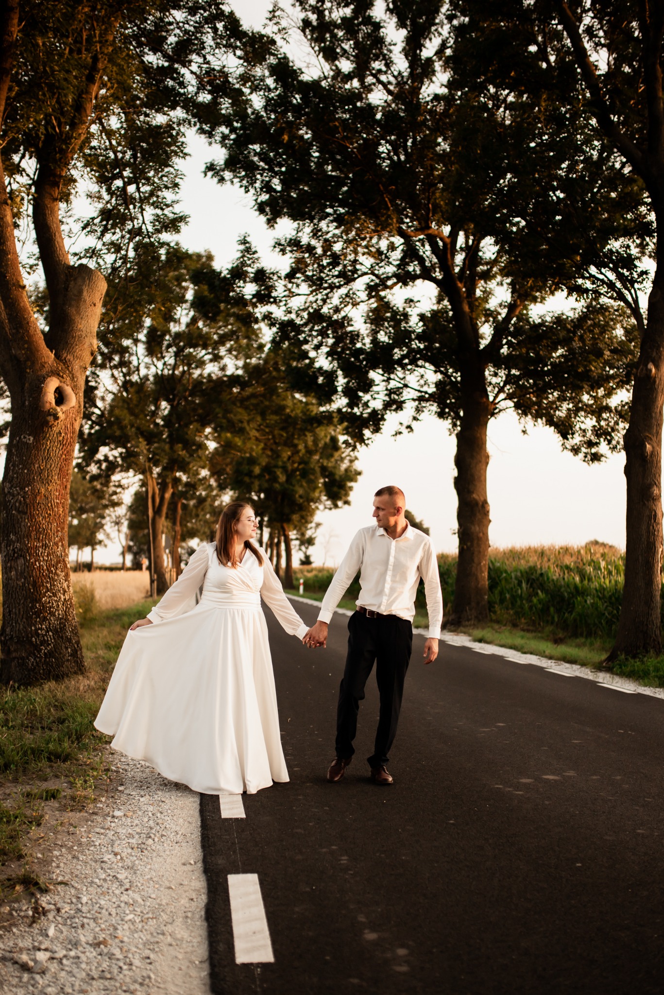 Para młoda idzie trzymając się za ręce po asfaltowej drodze otoczonej drzewami w ciepłym, zachodzącym słońcu. Kobieta w zwiewnej, białej sukni, mężczyzna w białej koszuli.