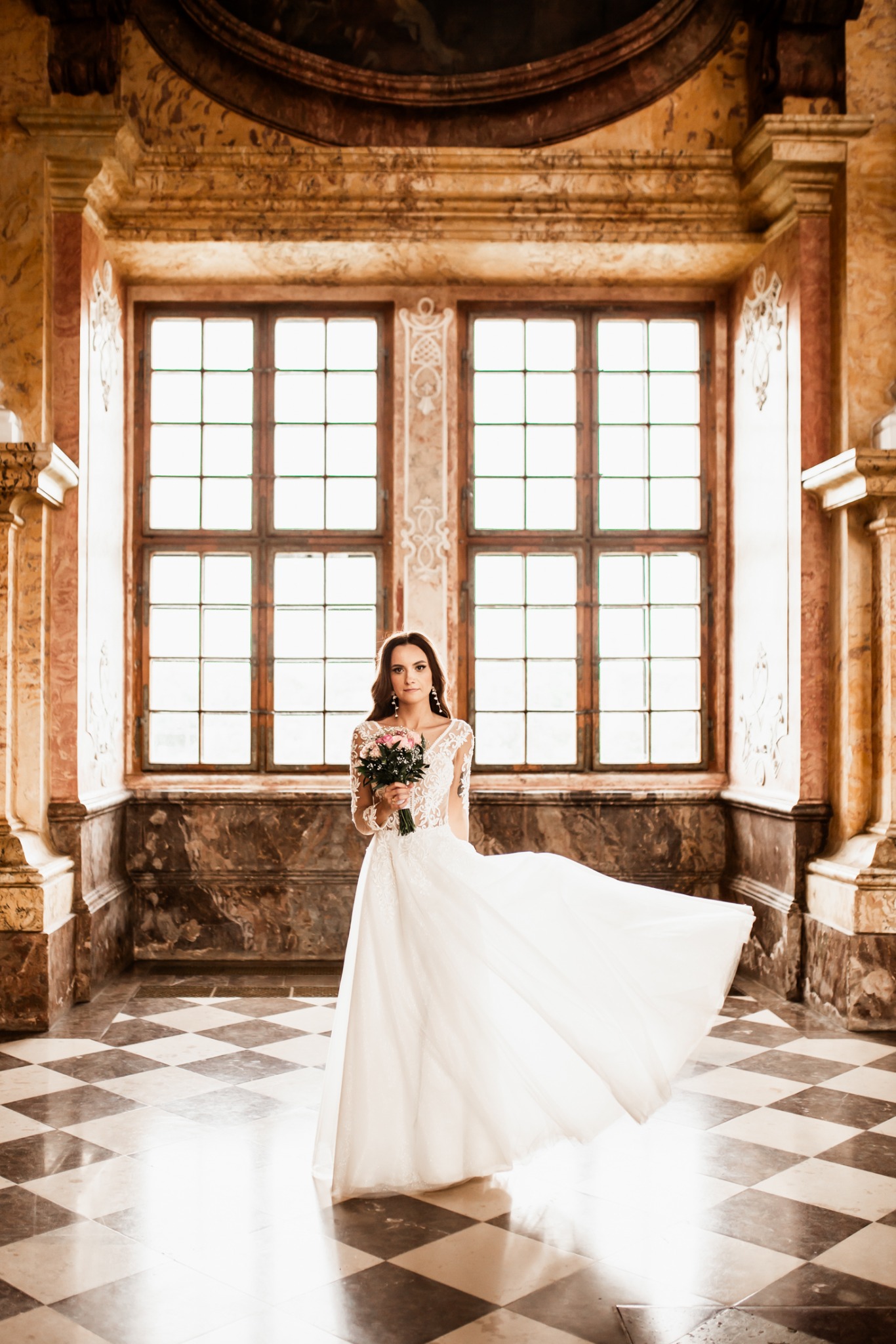 Panna młoda w koronkowej sukni z bukietem pozuje na tle marmurowych ścian i okien w stylu retro. Suknia rozwiana, podłoga w szachownice, ciepłe światło.