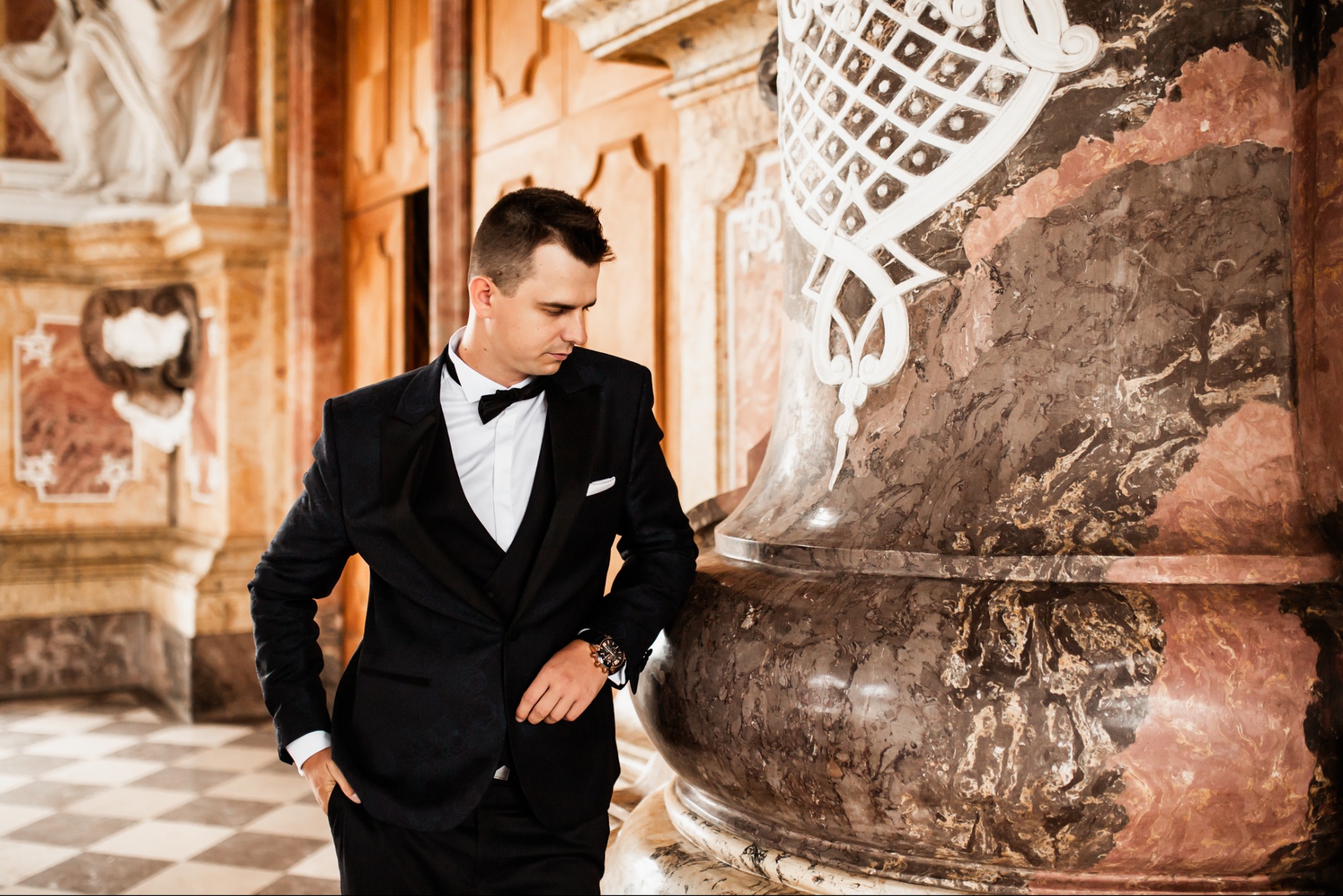 Elegancki mężczyzna w smokingu pozuje przy marmurowej kolumnie z dekoracyjnymi ornamentami w luksusowym wnętrzu. Subtelne światło podkreśla detale stroju.