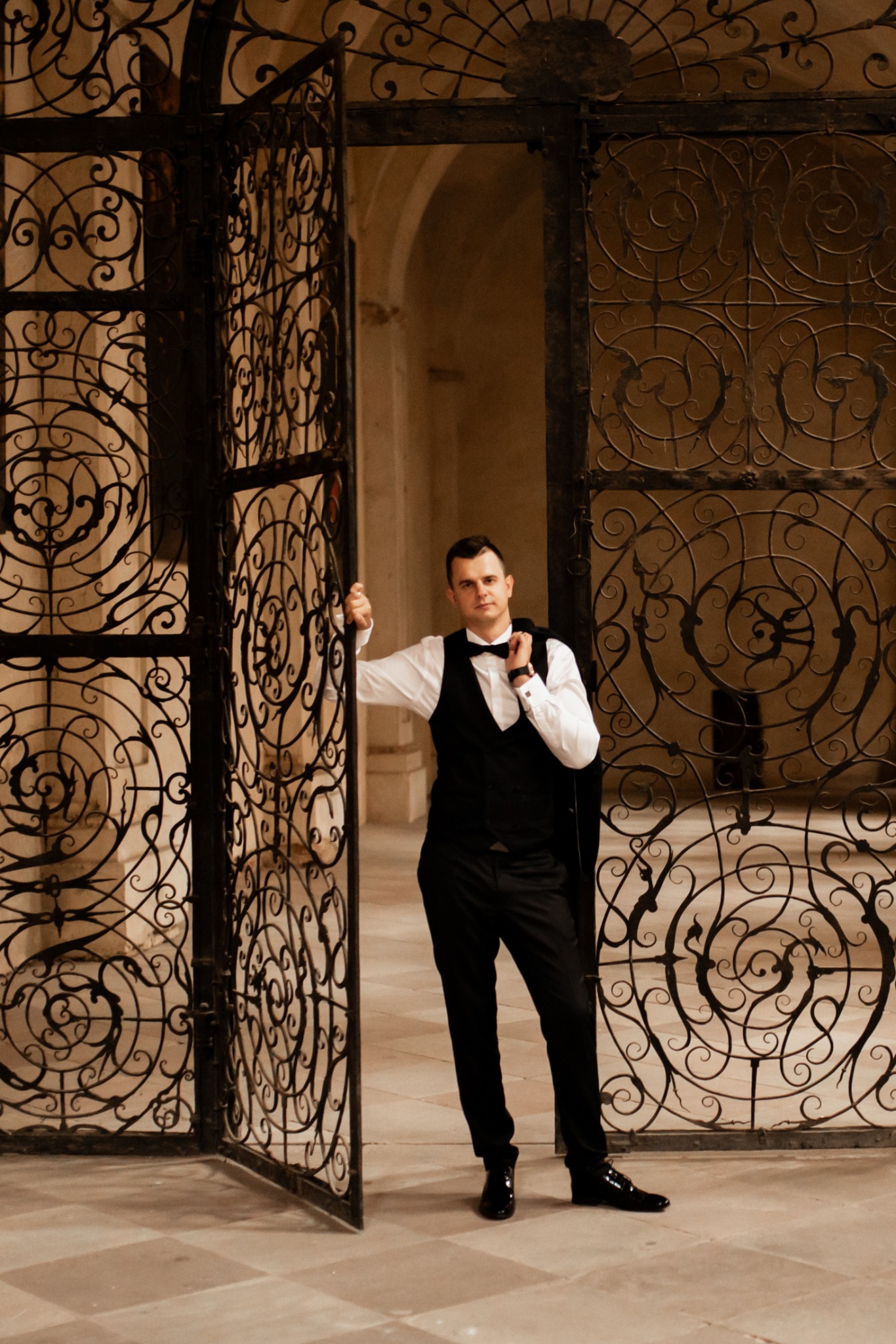 Elegancki mężczyzna w czarnej kamizelce i muszce pozuje przy otwartej, zdobionej metalowej bramie w ciepłym oświetleniu wnętrza. Ujęcie portretowe.