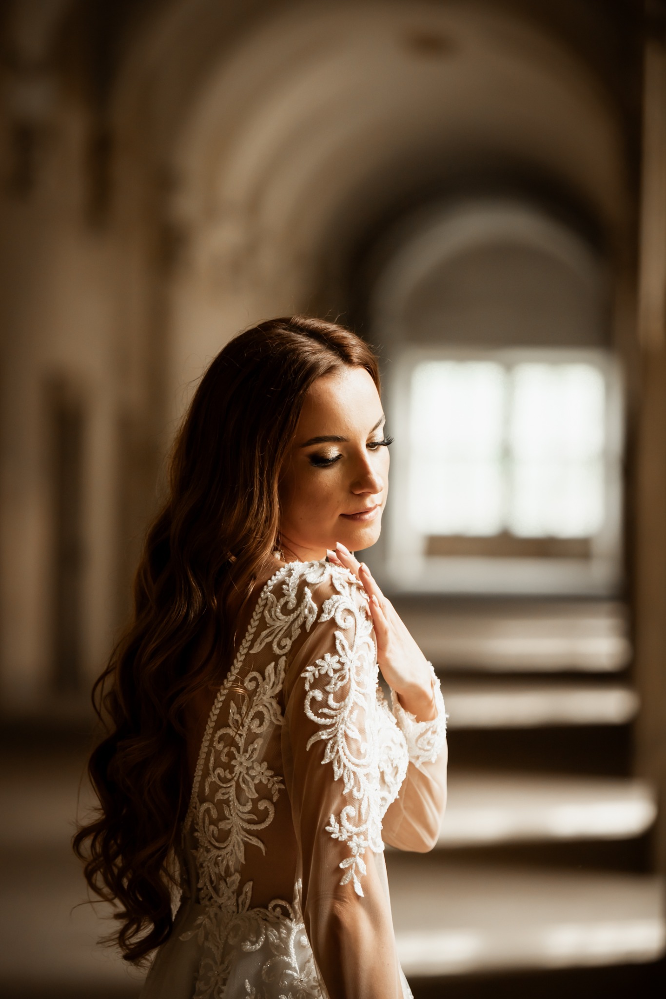 Portret kobiety w koronkowej sukni, stojącej w korytarzu z łukowatym sufitem. Delikatne światło podkreśla detale sukni i długie, brązowe włosy modelki.