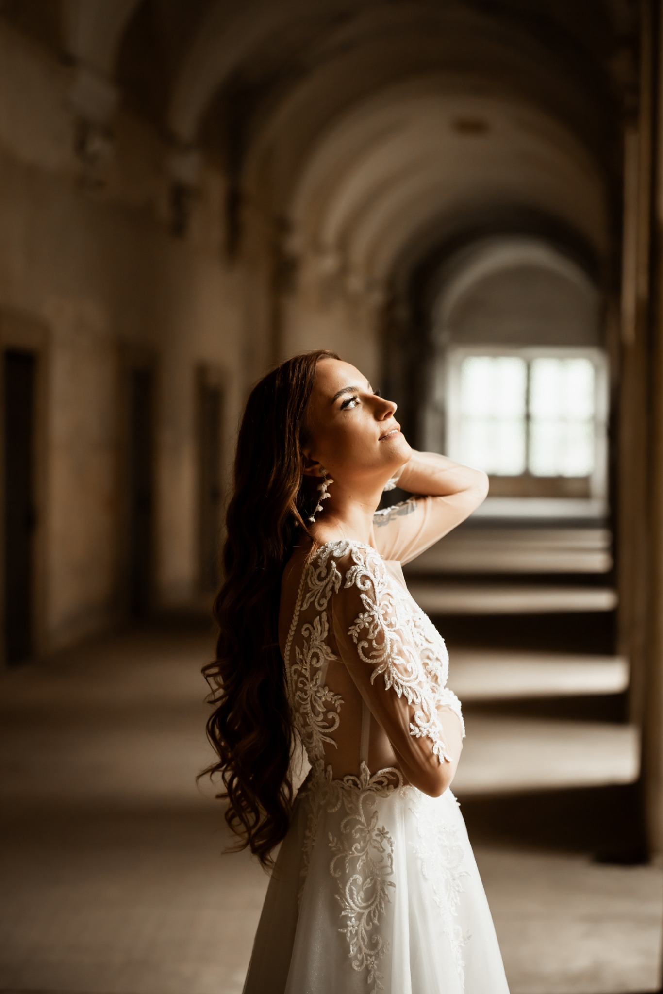 Panna młoda w koronkowej sukni spogląda w górę w korytarzu z łukami. Długie, brązowe włosy i subtelny makijaż. Delikatne kolczyki.