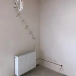 Biały grzejnik panelowy zamontowany na ścianie, z widoczną instalacją przewodów w peszlu, przymocowaną do ściany. Surowy stan wykończenia pomieszczenia.