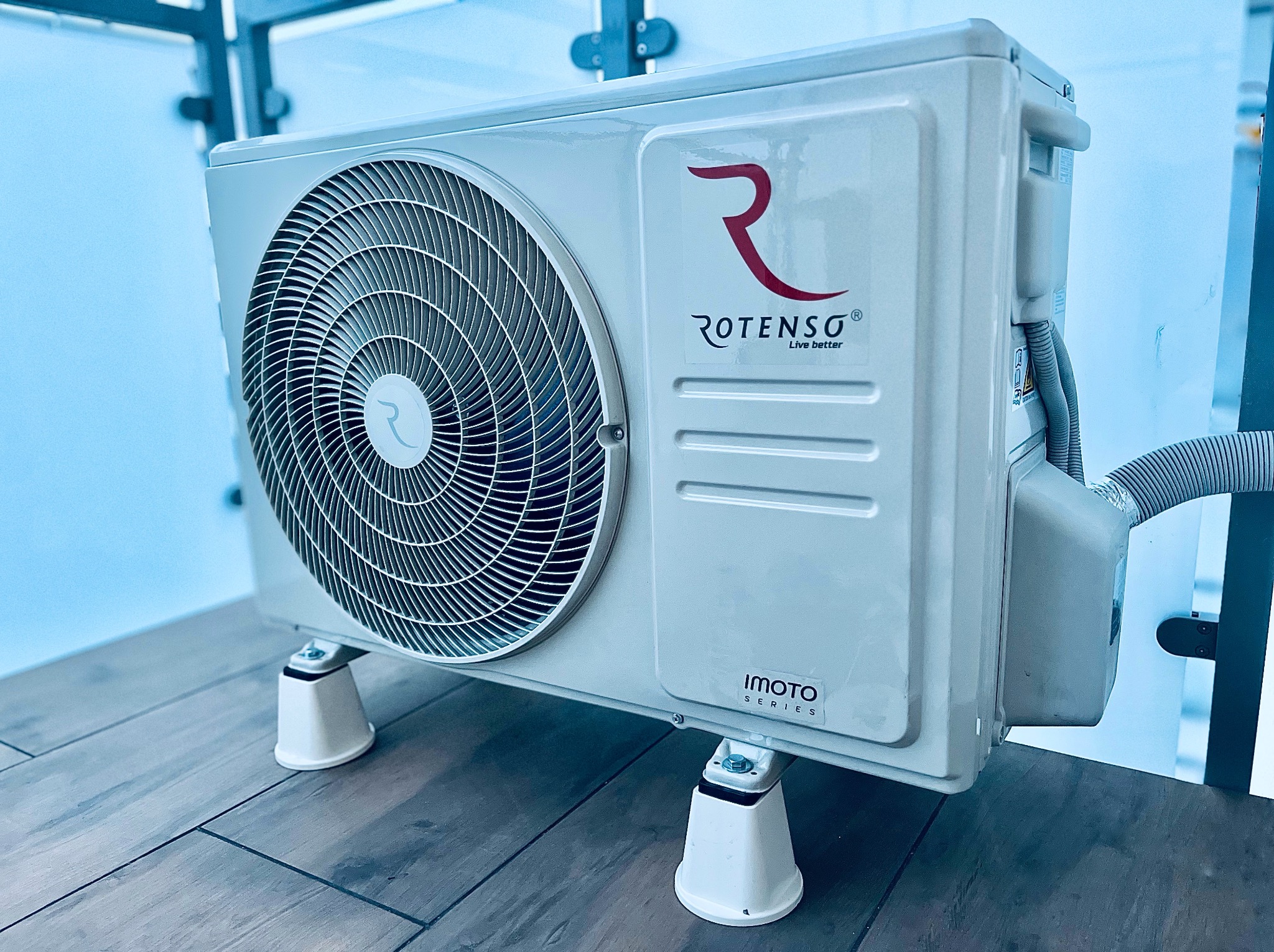 Biała, zewnętrzna jednostka klimatyzacji Rotenso IMOTO Series na czterech nóżkach, ustawiona na drewnianym blacie, z widocznym wężem odprowadzającym.