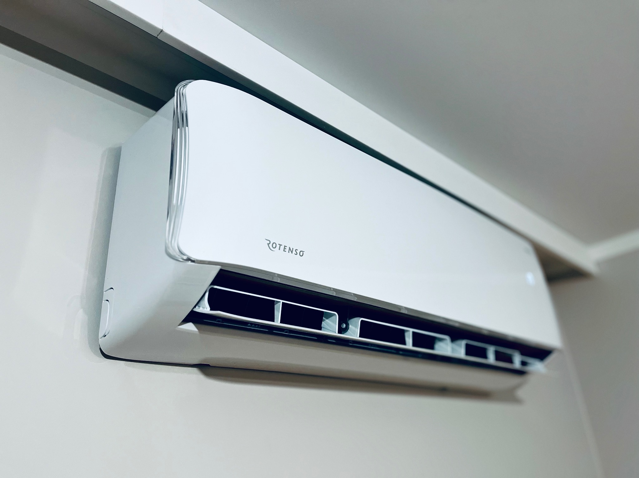 Biała klimatyzacja Rotenso, zamontowana na ścianie w jasnym wnętrzu. Widoczny detal srebrnego wykończenia i żaluzje wylotowe powietrza. Minimalistyczny design.