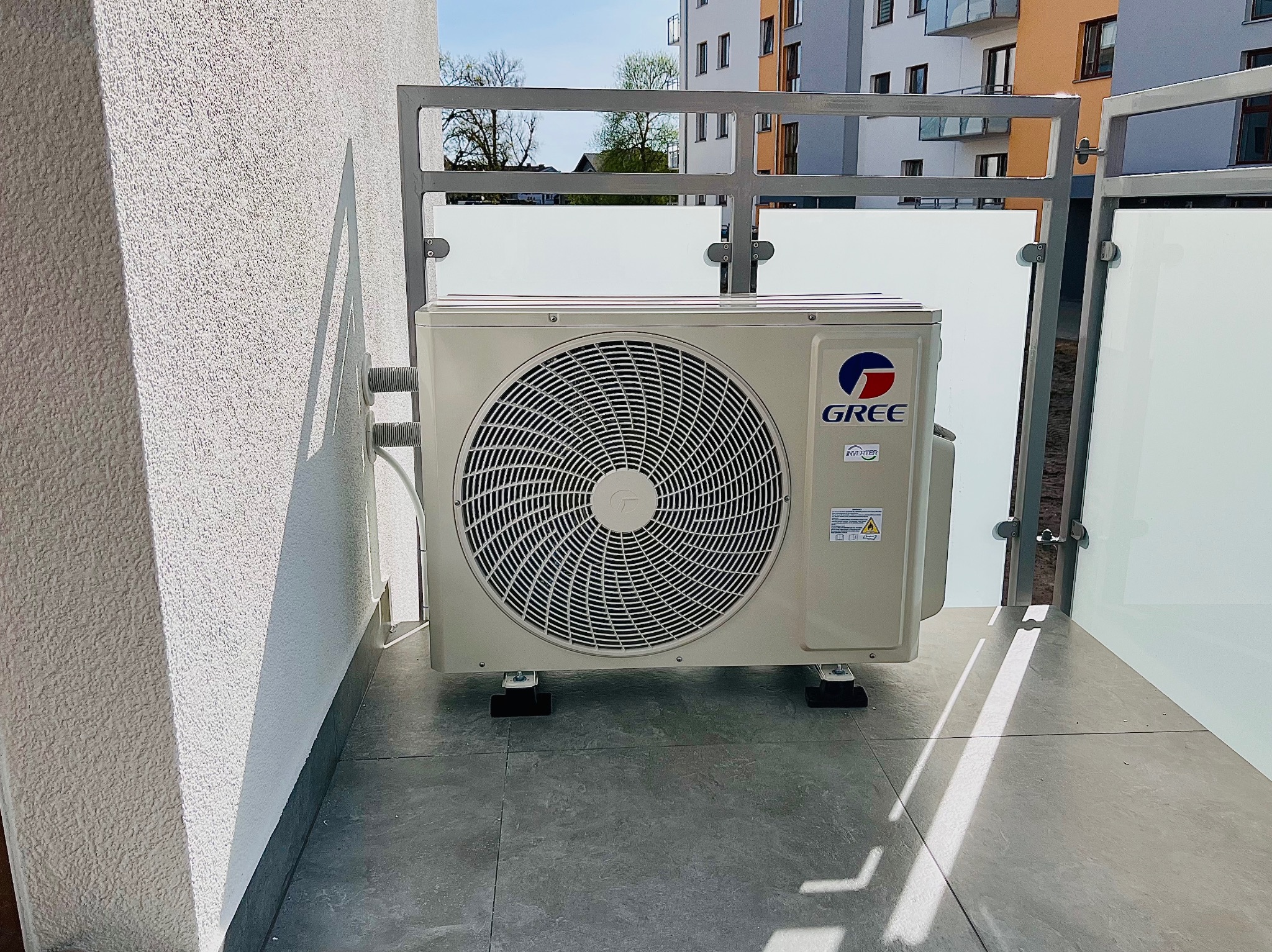 Klimatyzator Gree na balkonie, zamontowany na specjalnych podstawkach. Widoczne elementy montażowe i fragment elewacji budynku w tle. Ujęcie z poziomu podłogi.