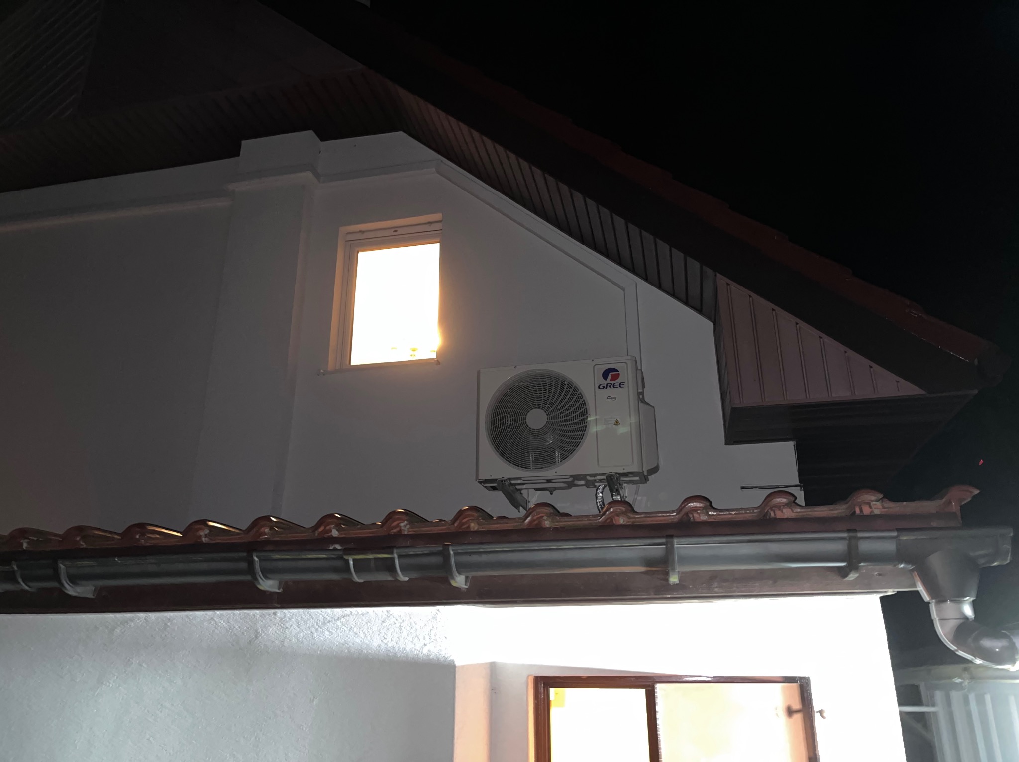 Biała jednostka zewnętrzna klimatyzacji Gree zamontowana na ścianie budynku z brązową dachówką, widoczna pod oknem z jasnym światłem w tle.