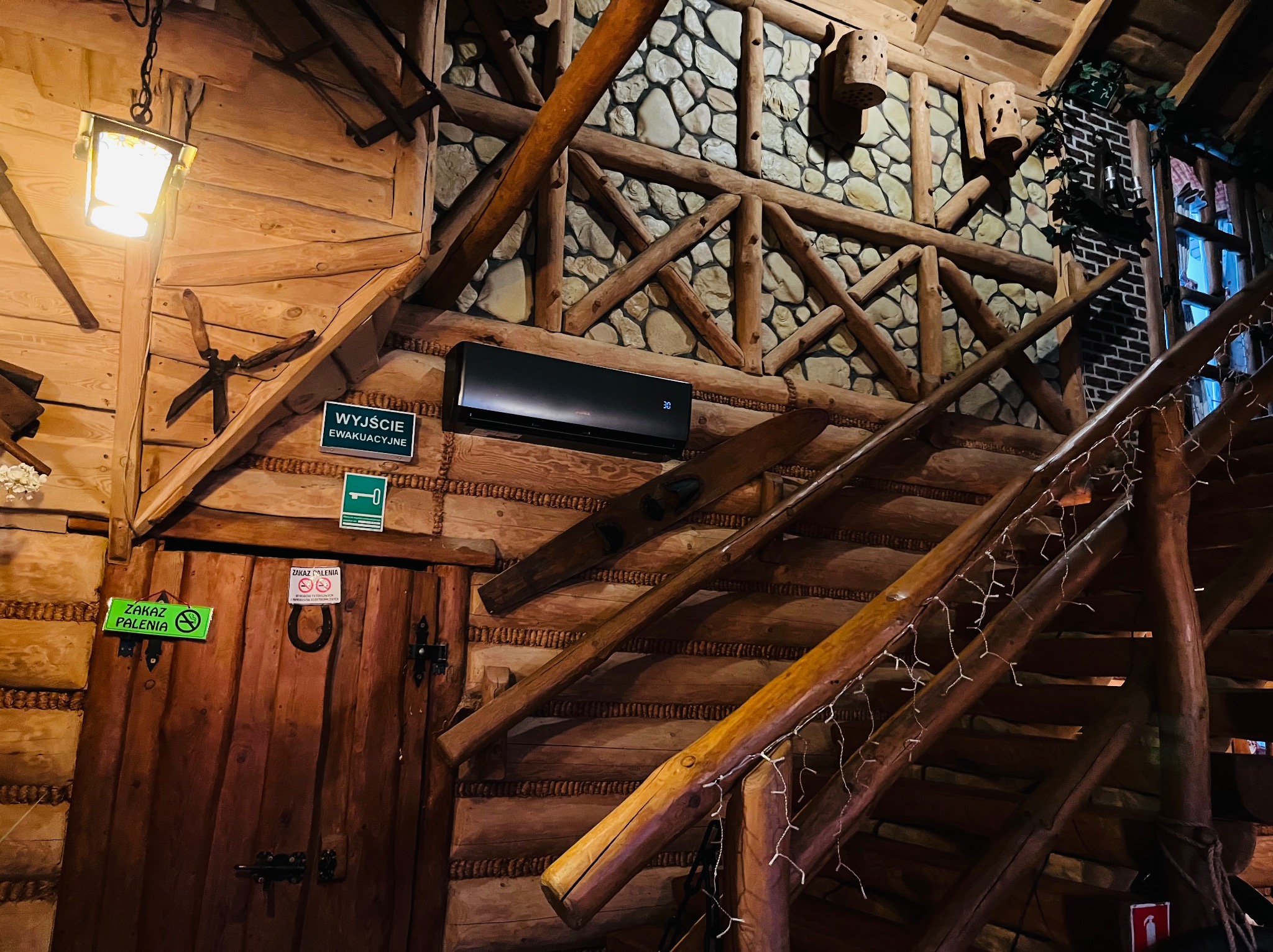 Klimatyzator ścienny w rustykalnym wnętrzu z drewnianymi schodami i elementami kamiennymi. Widoczne znaki informacyjne i dekoracje.
