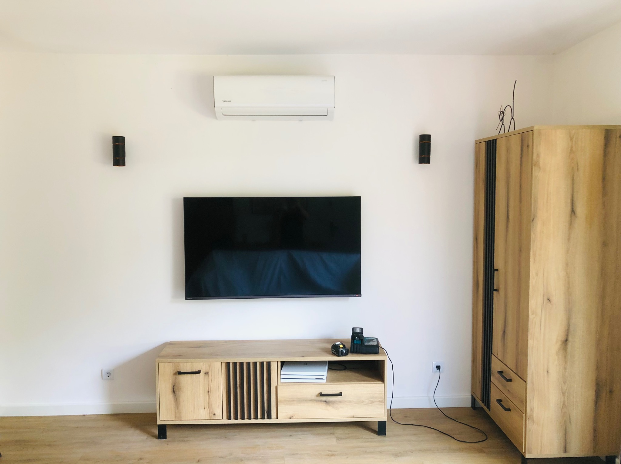 Nowoczesny salon z klimatyzatorem na ścianie, telewizorem, drewnianą szafką RTV i szafą. Minimalistyczny design, ciepłe oświetlenie, drewniana podłoga.