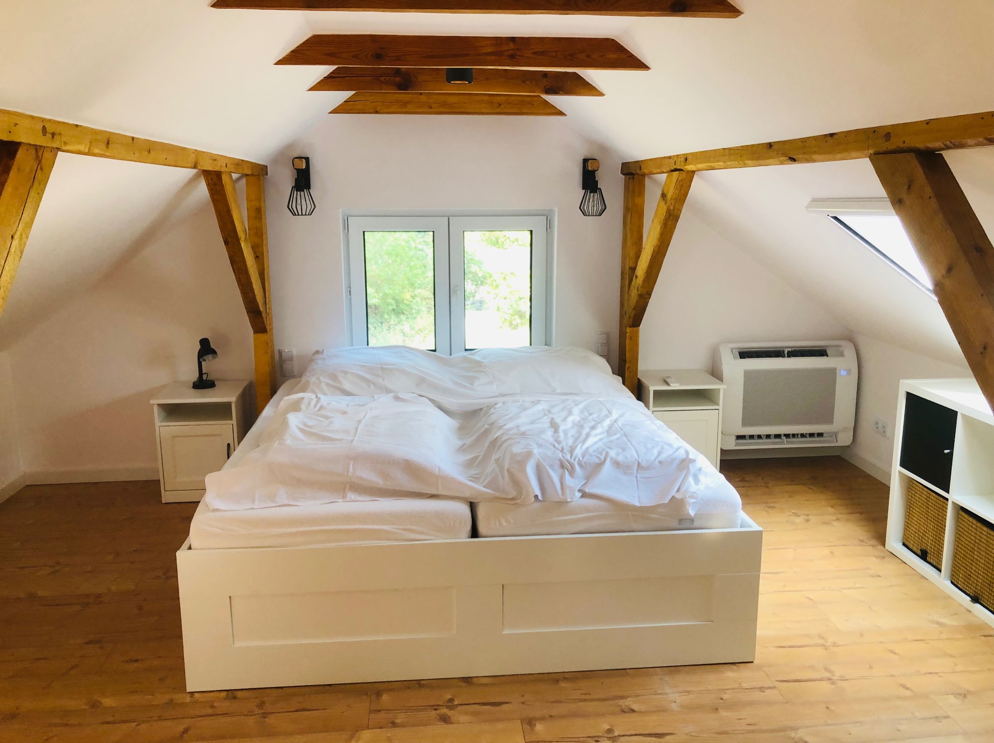 Jasna sypialnia na poddaszu z klimatyzatorem ściennym, drewnianymi belkami i białymi meblami. Widok na okno i skosy dachu.