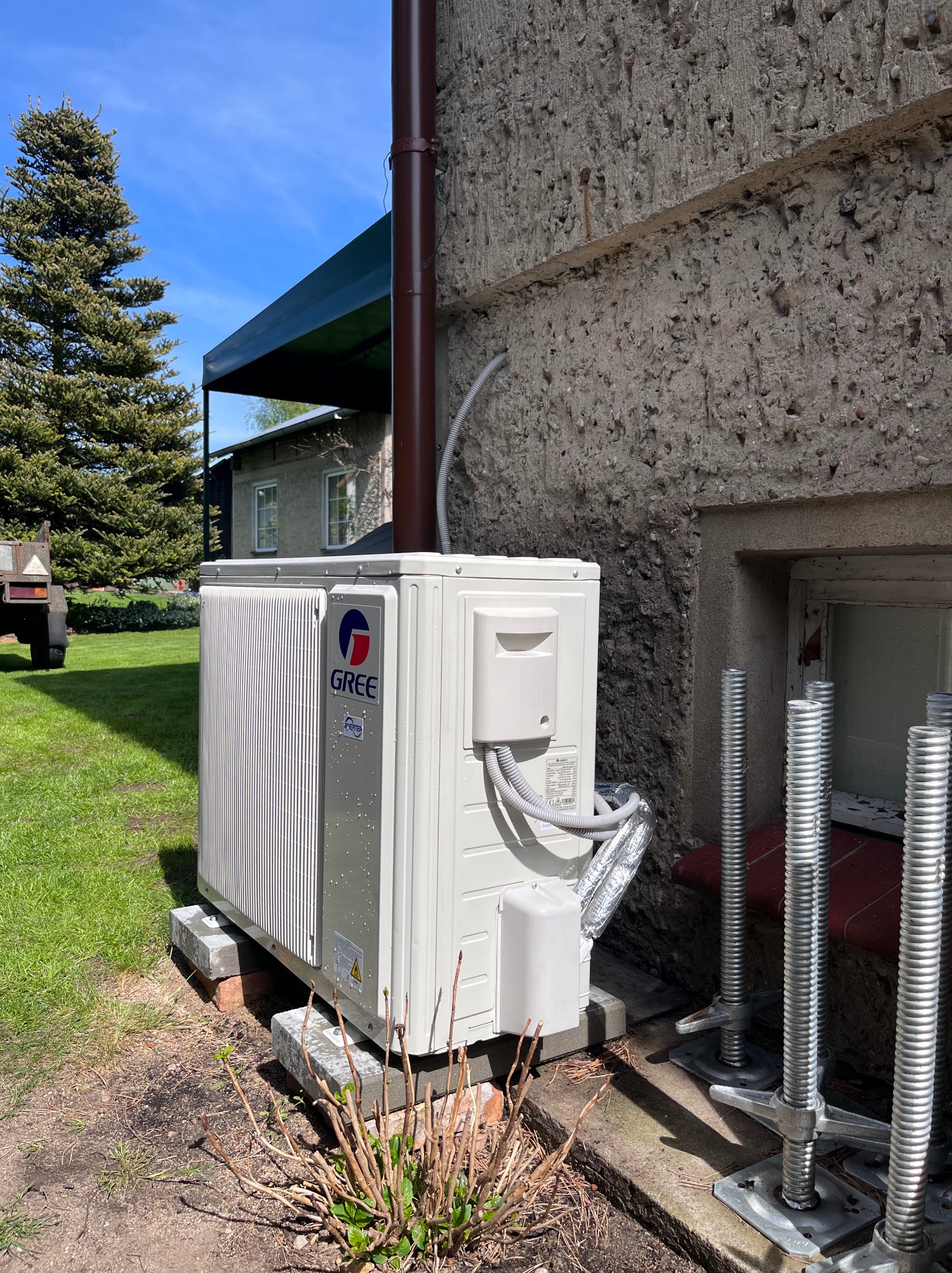 Biała jednostka zewnętrzna klimatyzacji Gree na betonowych podstawach przy szarej ścianie budynku, z widocznymi elementami instalacji i wspornikami montażowymi.