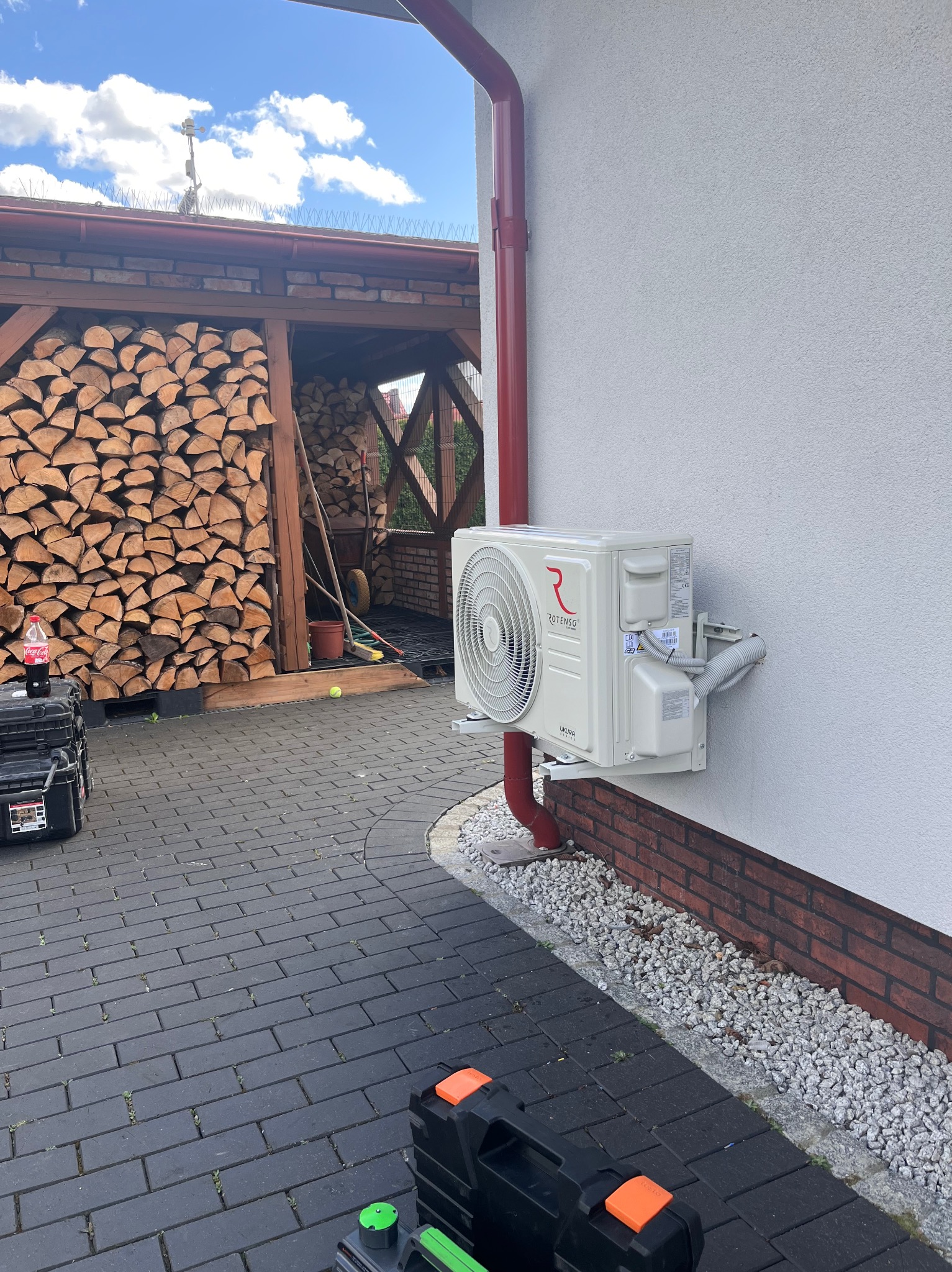Biała jednostka zewnętrzna klimatyzacji Rotenso, zamontowana na ścianie budynku z czerwoną rynną. Na pierwszym planie narzędzia instalacyjne, w tle drewno opałowe.