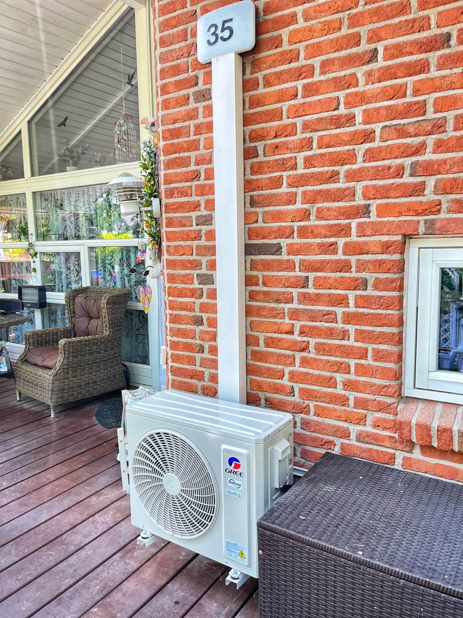 Biała jednostka zewnętrzna klimatyzacji GREE zamontowana na drewnianym tarasie przy ceglanej ścianie budynku z numerem 35. Widoczne elementy instalacji.