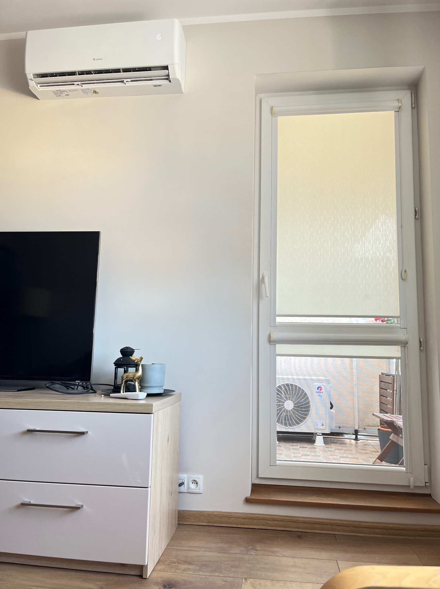 Biały klimatyzator Gree na ścianie w pokoju, widoczny przez okno agregat zewnętrzny. Nowoczesny design, minimalistyczne wnętrze.