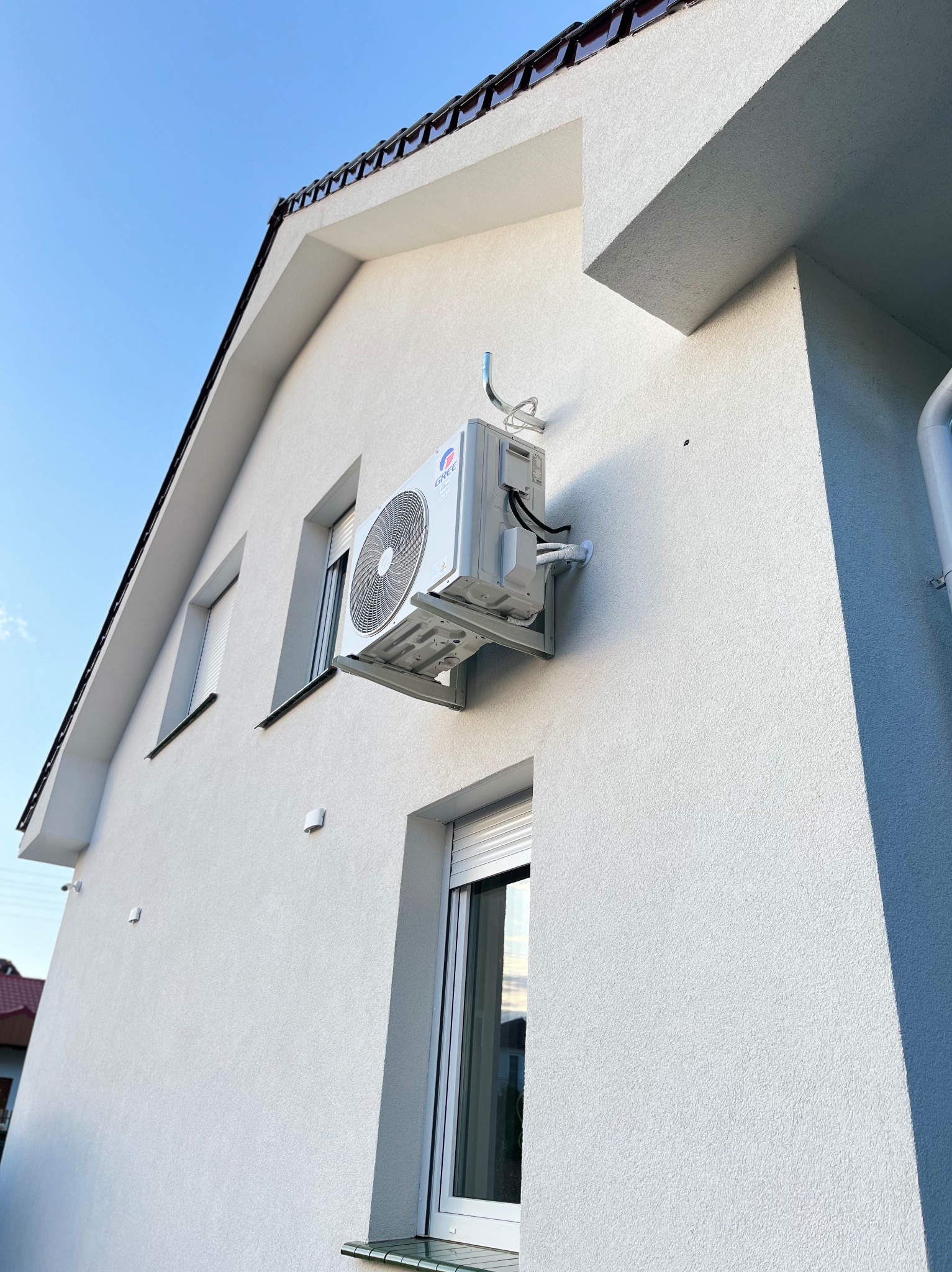 Biała jednostka zewnętrzna klimatyzacji Gree zamontowana na ścianie domu z oknami i brązowym dachem, widok z dołu na błękitne niebo.