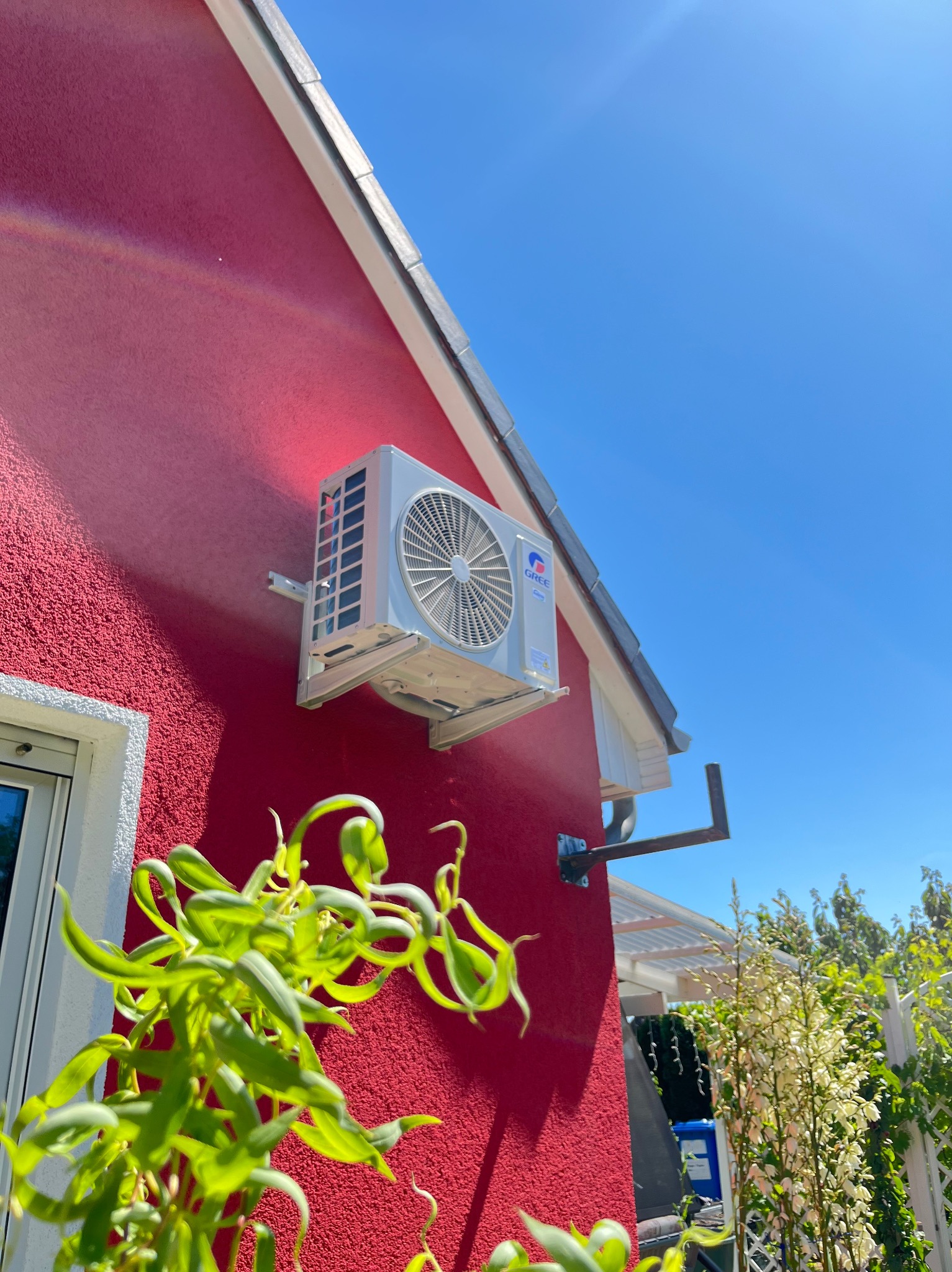 Biała jednostka zewnętrzna klimatyzacji Gree zamontowana na czerwonej ścianie domu, widok z dołu, z zielonymi roślinami na pierwszym planie i błękitnym niebem w tle.