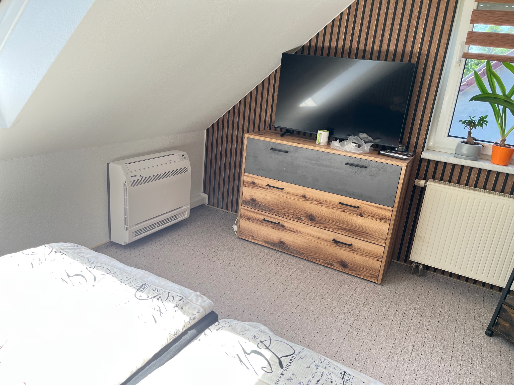 Biała klimatyzacja podłogowa Gree w pokoju na poddaszu, obok komody z telewizorem i grzejnika pod oknem. Ściana w dekoracyjnych panelach.