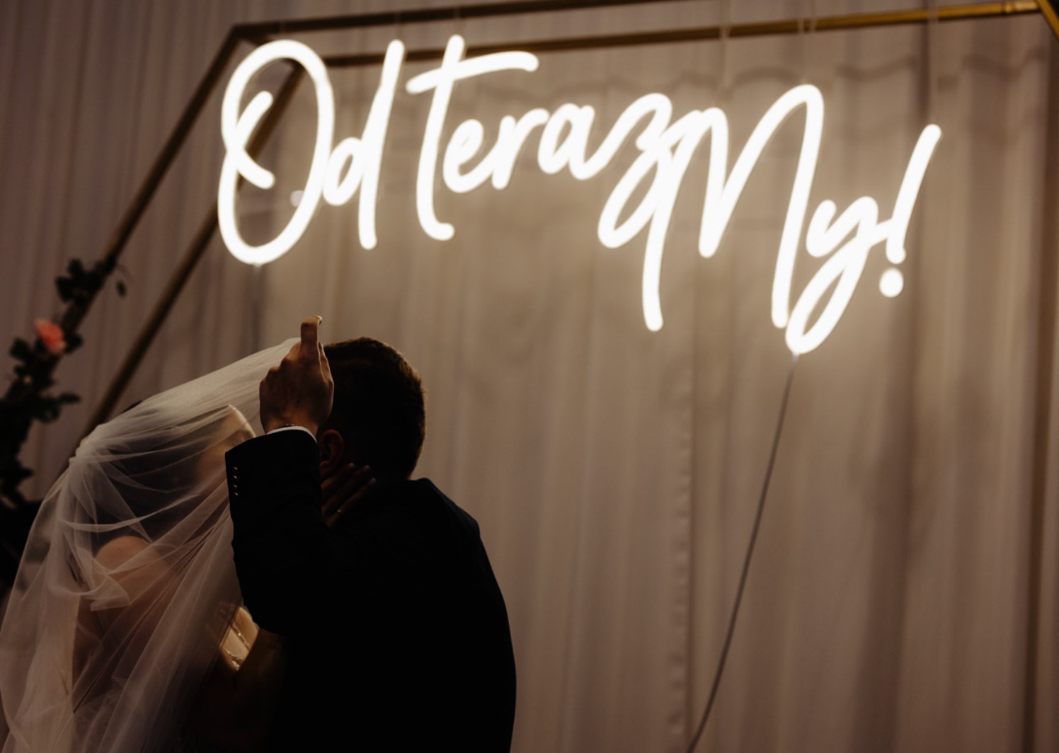 Pocałunek pary młodej pod neonem 'Od teraz My!' w geometrycznej ramie. Pan Młody w ciemnym garniturze, Panna Młoda pod welonem. Romantyczna sceneria ślubna.