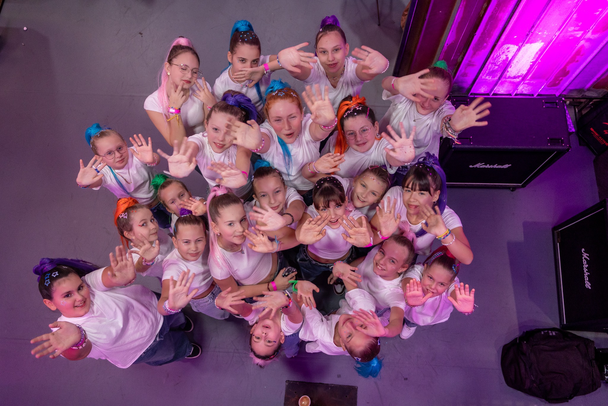 Grupowe ujęcie z góry: zespół taneczny dziecięcy w białych koszulkach, z kolorowymi włosami i brokatem, pozuje z uśmiechem na tle sprzętu nagłośnieniowego.