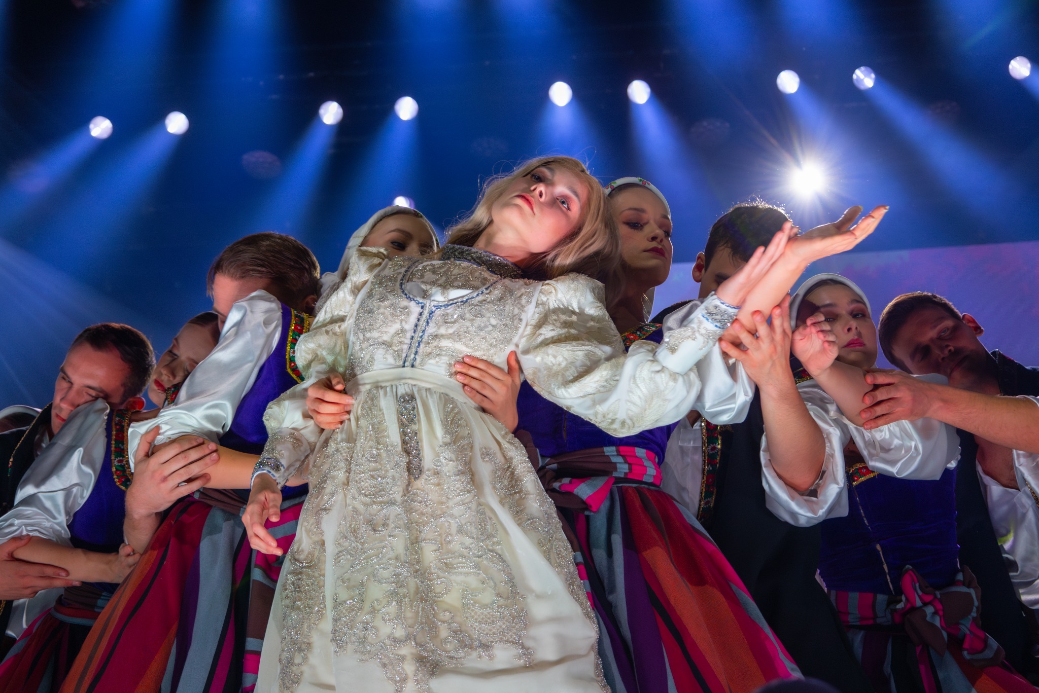 Grupa tancerzy w barwnych strojach ludowych pod światłami sceny, w dynamicznej pozie podczas występu. Centralna postać w bogato zdobionej, białej sukni.