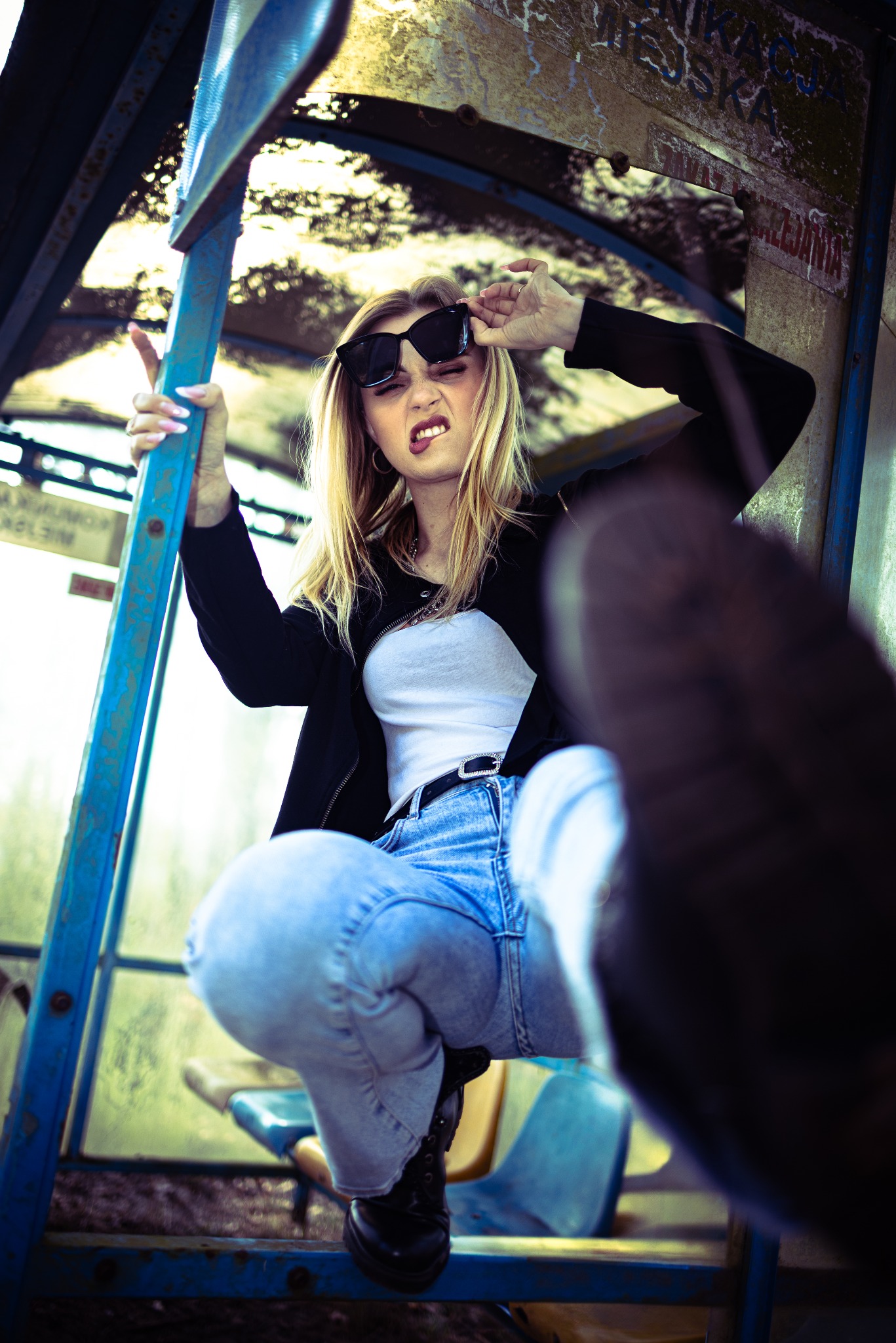 Ekspresyjna blondynka w dżinsach i okularach pozuje na przystanku autobusowym, ukazując swój charakterystyczny styl i niekonwencjonalne podejście do mody.