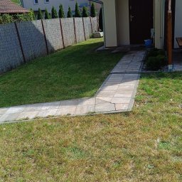 Paula Zygmunt - Świeżo skoszony trawnik z wybrukowaną ścieżką w słoneczny dzień. Widoczny budynek z brązowymi drzwiami, zielony żywopłot i częściowo widoczne drzewa.