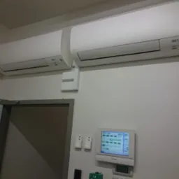 Dwa klimatyzatory Daikin zamontowane na białej ścianie, obok panel sterowania i drzwi. Widoczne elementy instalacji elektrycznej i sterowania.