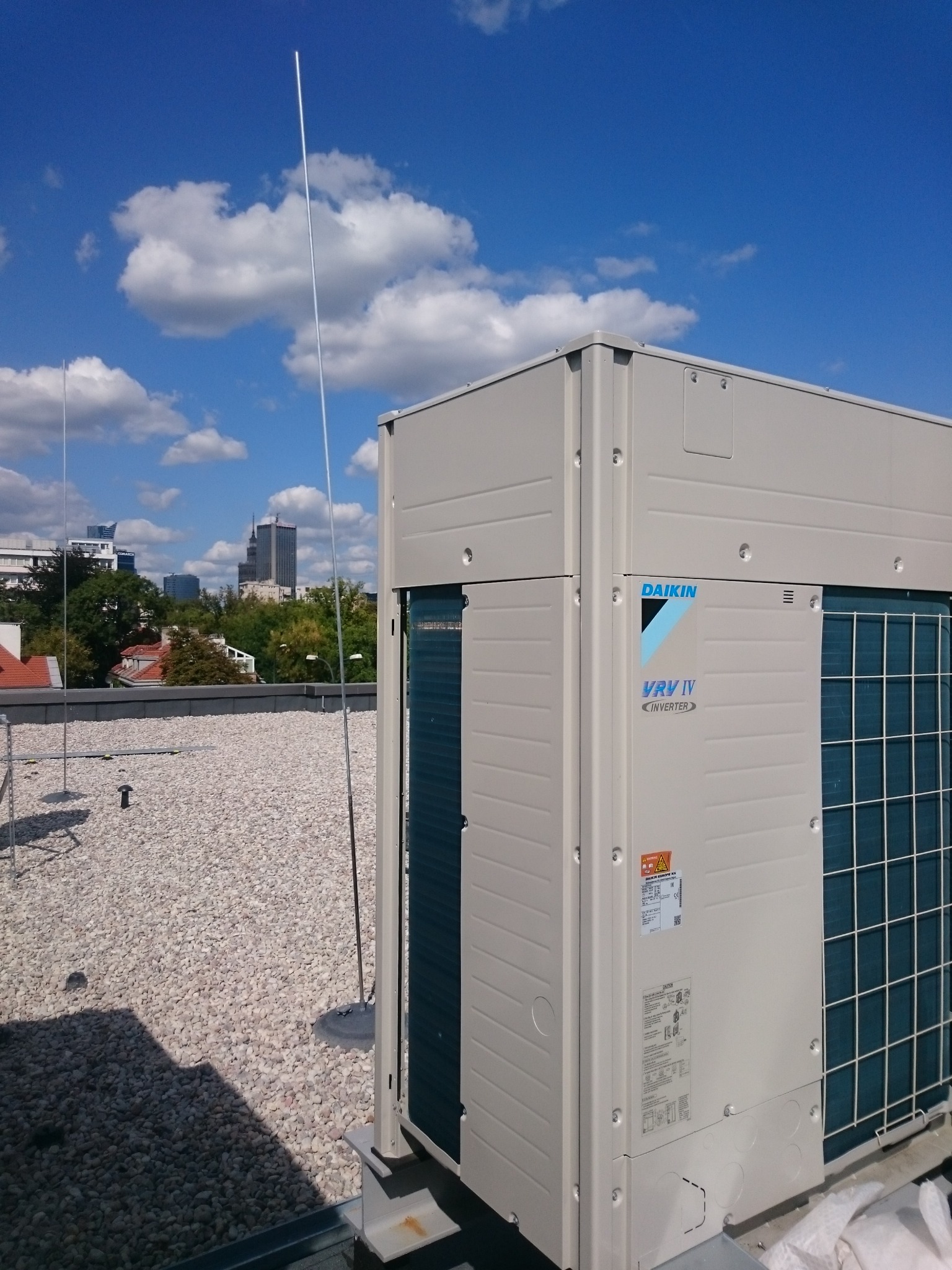 Klimatyzator Daikin VRV IV Inverter na dachu budynku, z widokiem na panoramę miasta w tle i niebieskie niebo z chmurami.
