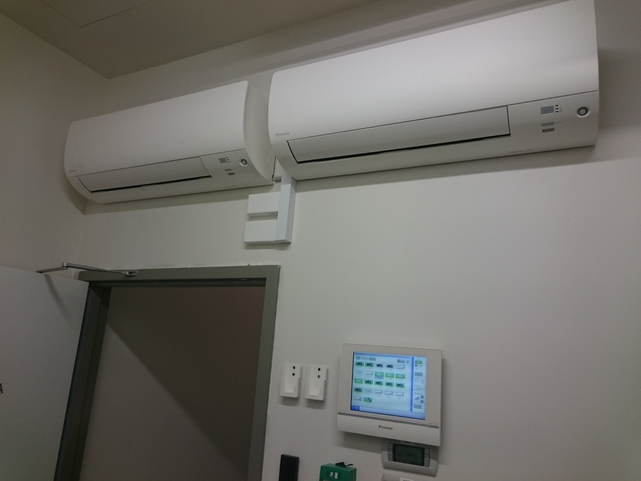 Dwa klimatyzatory Daikin zamontowane na białej ścianie, obok panel sterowania i drzwi. Widoczne elementy instalacji elektrycznej i sterowania.