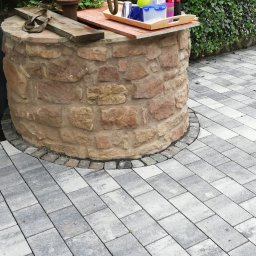 MARGELS SPÓŁKA Z OGRANICZONĄ ODPOWIEDZIALNOŚCIĄ - Studnia z kamienia otoczona szarą kostką brukową, schody z kostki prowadzą w dół. Zielona ściana z bluszczu w tle.